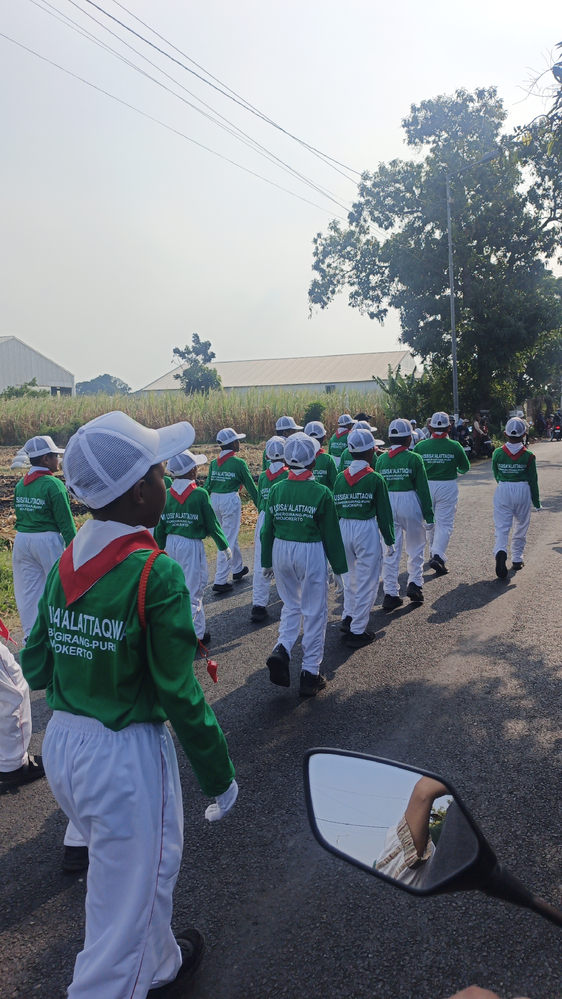 A group of children in matching green long-sleeved shirts and white pants with red trim are marching in a procession down an asphalt road. They are wearing white caps. Some of the children have red scarves around their necks. The shirts have text printed on the back. The environment appears to be semi-rural, with fields and buildings visible in the background. There are power lines overhead and a tree on the right side of the image. A motorcycle mirror in the foreground reflects a person's legs. The text on the shirts appears to be in Indonesian, potentially indicating a school or organization name and location such as "SISA'ALATTAQWA", "B'GIRANG-PURI", and "MOJOKERTO". The scene is outdoors in daylight.