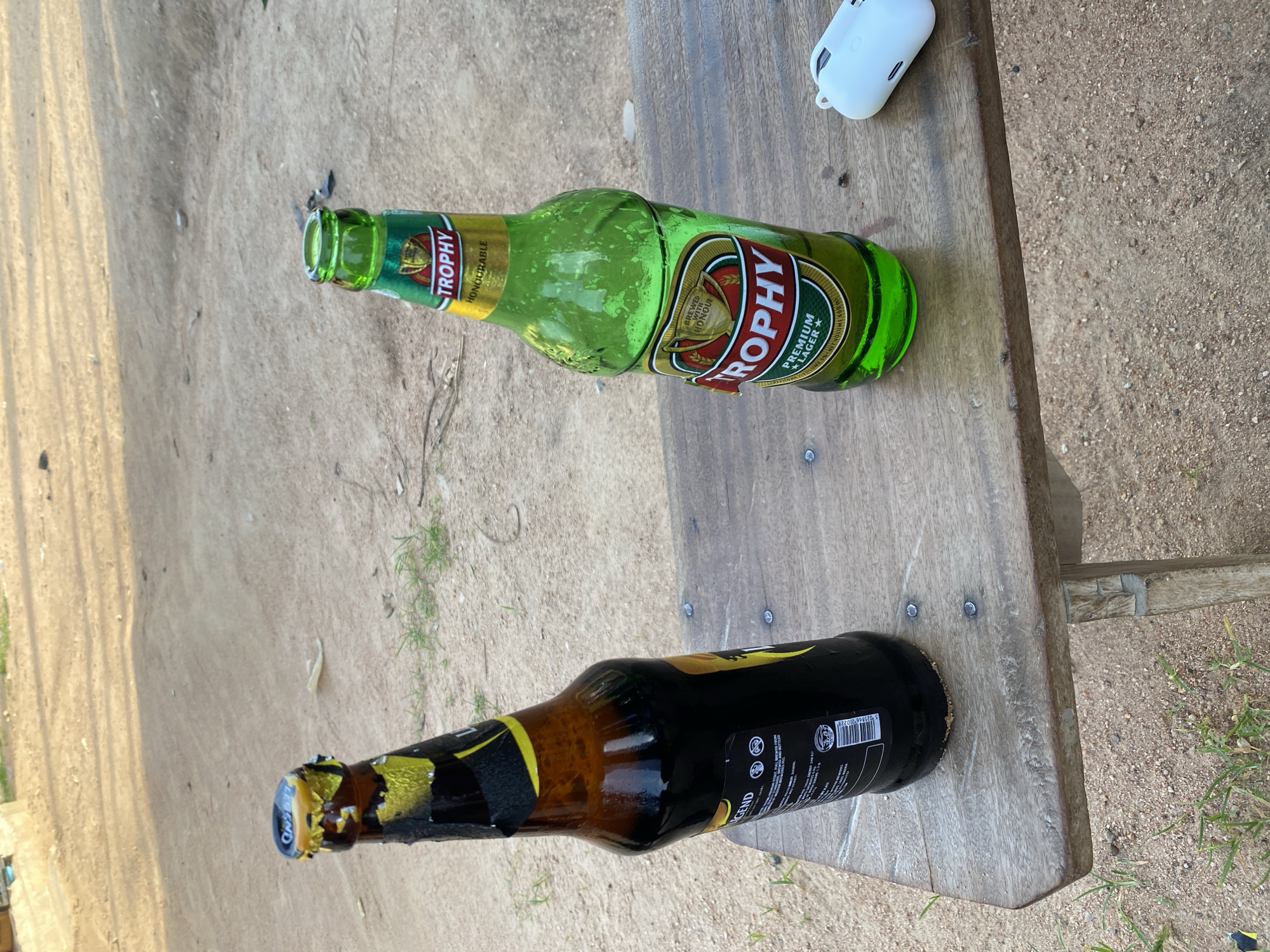 Two bottles of beer, one dark brown and one green, sit on a weathered wooden table outdoors. The dark brown bottle, labeled "LEGEND," has a partially removed gold foil cap. The green bottle, labeled "TROPHY," has a prominent red and gold label with "PREMIUM LAGER" printed on it. A white wireless earbud case is to the right of the bottles. The background consists of sandy ground with sparse dry grass, suggesting an outdoor, possibly rural or less developed area. The lighting indicates it is daytime. There are no people visible in the image. The scene is likely in Howayi, Nigeria, given the context.