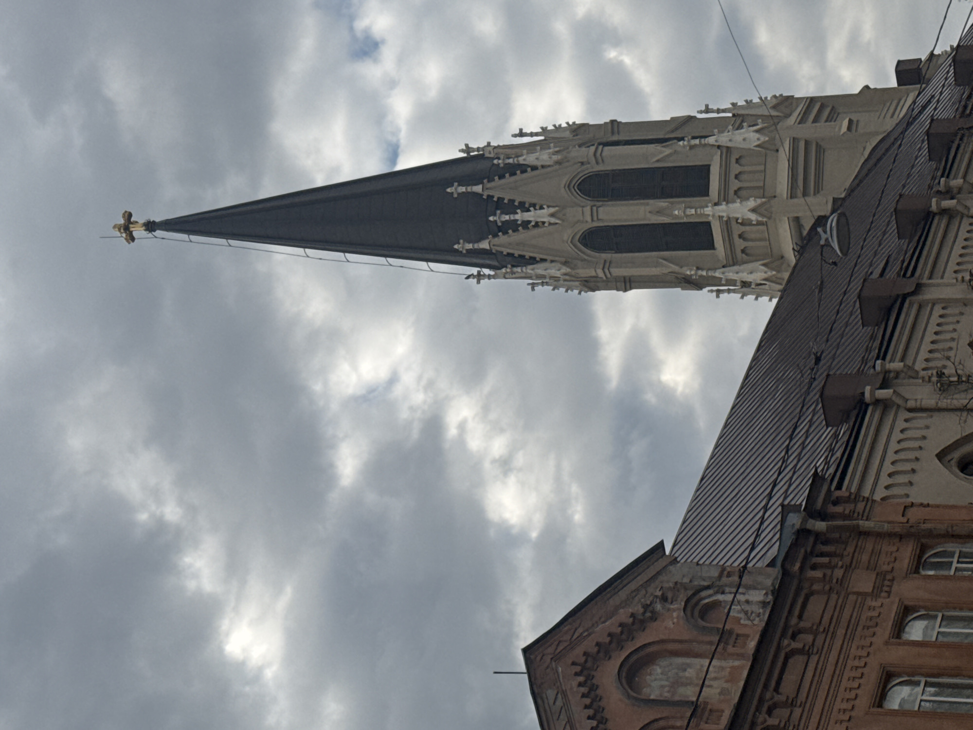 The image, rotated clockwise, features a tall, ornate Gothic-style church spire dominating the left side against a cloudy sky. The spire is dark and pointed, crowned with a small golden cross at its apex. Below the dark peak, the spire transitions into a lighter, possibly white or grey stone section, richly detailed with multiple smaller pinnacles, arched windows, and intricate carvings.
To the right, the roof and upper facade of an adjacent building are visible. This structure has a dark, possibly metal or tiled roof, and a facade in warm terracotta or brown tones. It features decorative elements, including arched window frames and patterned brickwork or plasterwork. Utility wires are seen traversing the space between the two buildings and against the sky.
The sky is heavily overcast, filled with various shades of grey clouds, though some brighter areas suggest diffused sunlight. The lighting indicates it is daytime. No people or specific activities are visible in the scene, which focuses primarily on the architectural details. No text is discernible in the image. The architectural styles are consistent with historic buildings found in cities like Saint Petersburg, Russia.