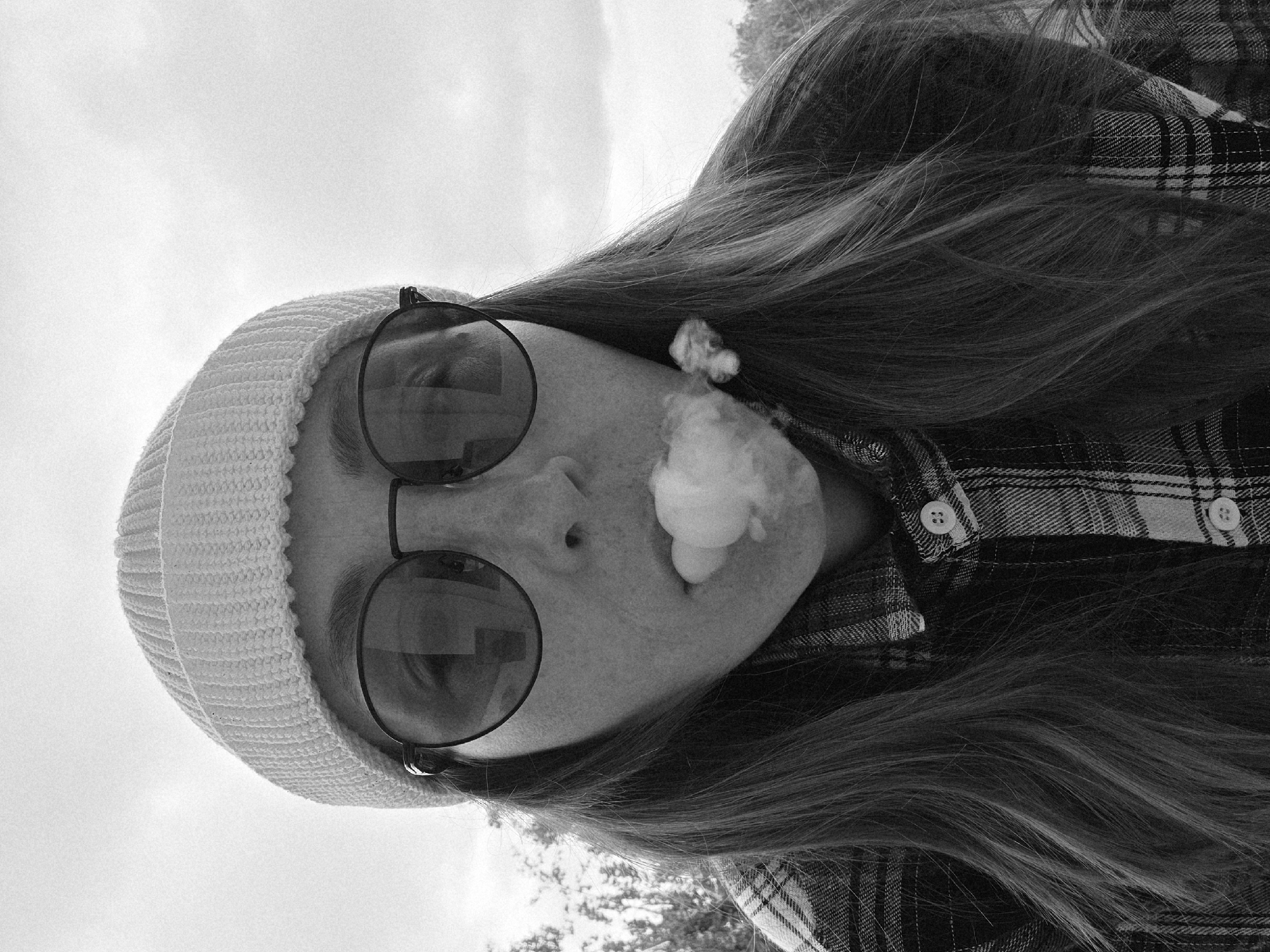 This black and white, grainy photograph, taken in Chaikovsky, Russia, captures a person from a low-angle perspective. The image is rotated 90 degrees counter-clockwise, showing the subject's head on the left and their body extending towards the right. The person is exhaling a visible cloud of vapor or smoke from their mouth, with some residual vapor near their ear.
They are wearing a light-colored, possibly white or cream, knit beanie and large, dark, round sunglasses that obscure their eyes. Their long, dark hair is prominent, flowing over their shoulder. The person is dressed in a plaid shirt, visible on the right side of the frame, with distinct checked patterns and buttons.
The background is predominantly a bright, seemingly overcast sky, indicating daylight conditions. Faint outlines of trees or foliage can be discerned on the far right, suggesting an outdoor setting. The black and white aesthetic, combined with the grainy texture, lends a candid or vintage feel to the photograph. No text is visible in the image.