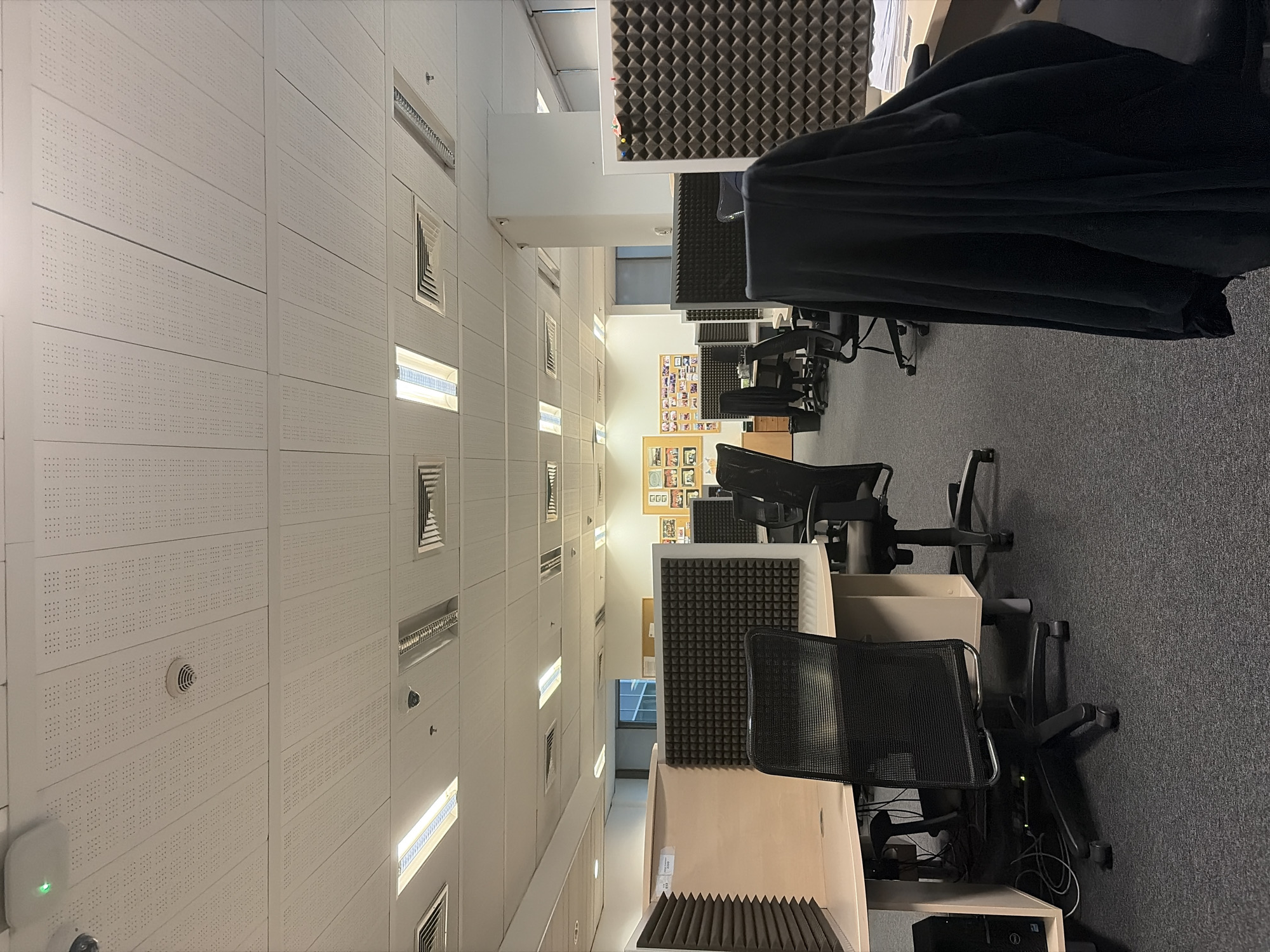 The image shows an office interior, likely a call center or similar workspace, in Sofia, Bulgaria. The layout features rows of workstations with acoustic panels, typical for reducing noise in open-plan environments. There are office chairs and computer equipment visible at the workstations. The walls and ceiling are primarily white, with integrated lighting fixtures and ventilation grilles. A corkboard displaying pictures and notes is visible further down the hallway. The scene is generally tidy, with an emphasis on functional office design. There are no people present in the immediate foreground, although distant figures may be obscured by the cubicle walls. The flooring appears to be gray carpet.