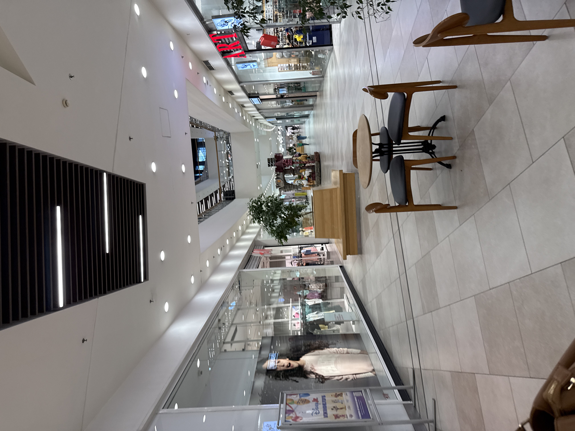 The image shows the interior of a shopping mall, likely in Stara Zagora, Bulgaria, based on the shop names visible. The foreground contains seating areas with round tables and modern chairs. Further into the frame, the mall corridor stretches into the distance, lined with shops behind glass facades. A mannequin in a storefront display is visible on the left. Shoppers walk through the corridor. Above, the ceiling features linear lighting and architectural elements.