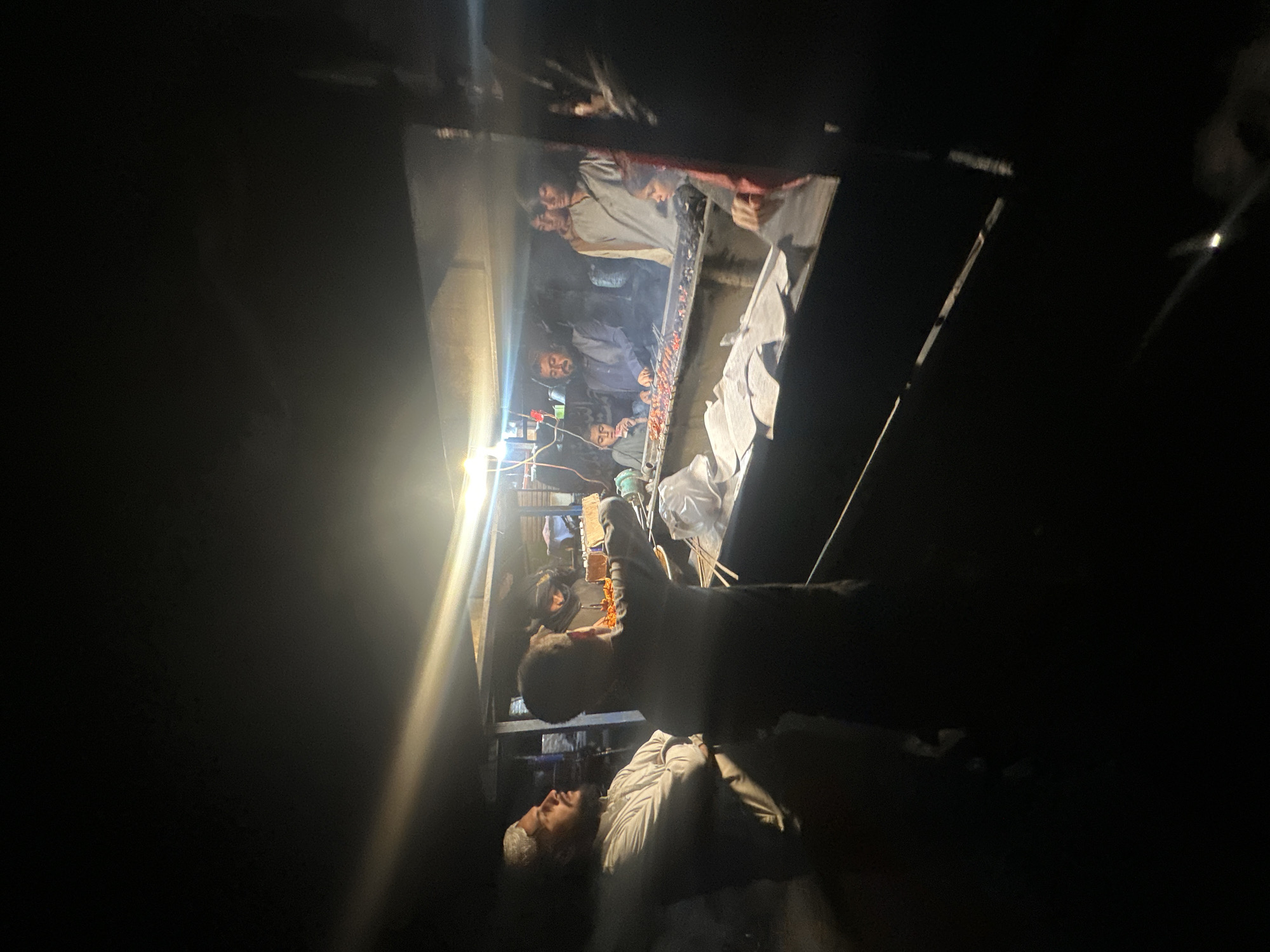 In Quetta City Tehsil, Pakistan, the image captures a dimly lit scene, with a group of men visible within what appears to be an elevated, enclosed space. Several men are gathered together in the elevated area, and their expressions and postures suggest they are interacting with something within the space. Below this elevated area, another man is visible. He is wearing a light-colored shirt and appears to be looking upwards towards the group of men above. The environment around the men is dark, adding to the overall subdued and somewhat mysterious feel of the image. The scene is focused on the interactions between the individuals and the illuminated space they occupy.