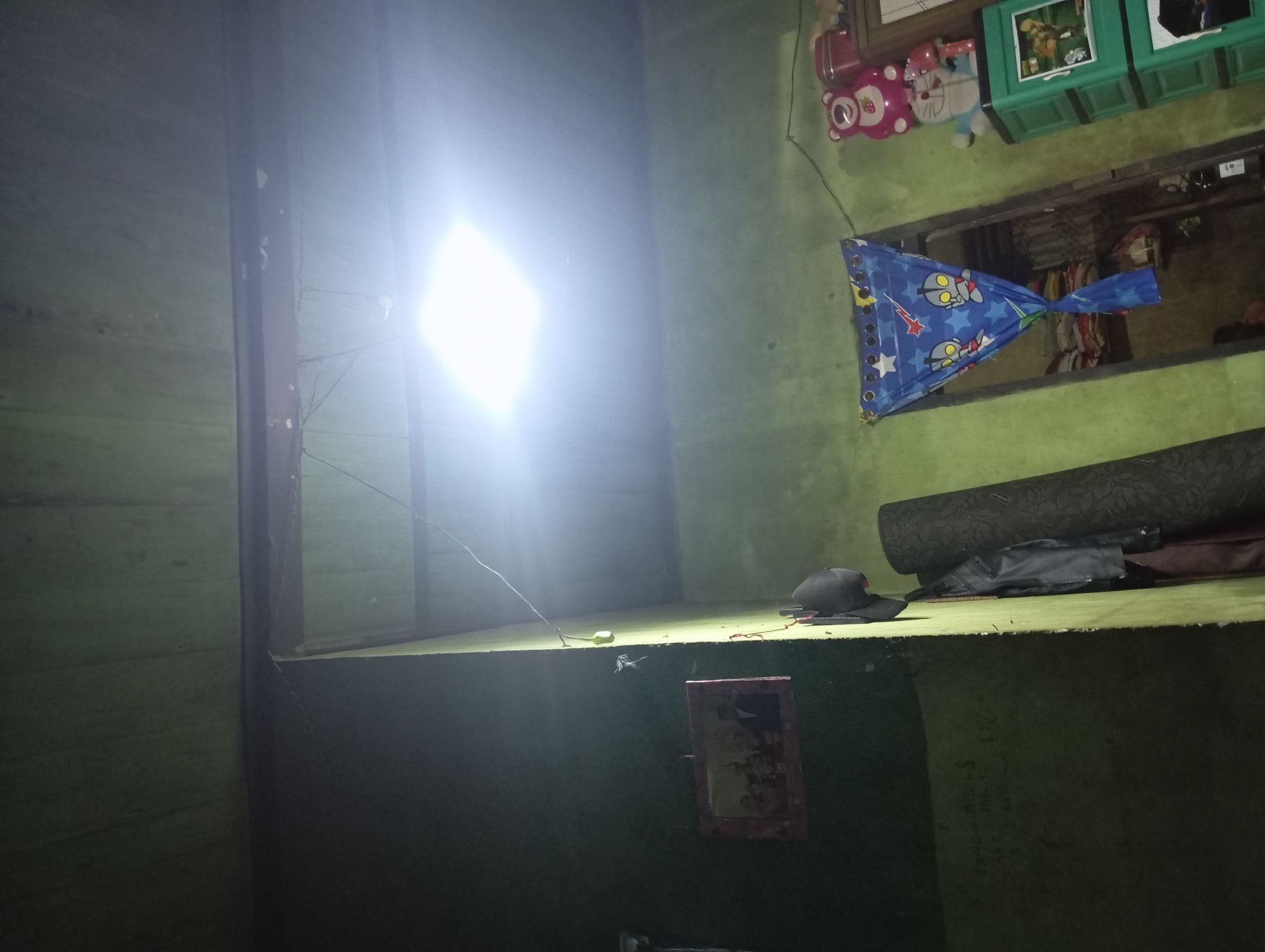 This is an indoor, eye-level, medium-shot photograph of a dimly lit room in Rejosari, Indonesia. The room appears to be a residential space with green painted walls and a wooden ceiling. A bright, square ceiling light illuminates the upper right portion of the scene, creating harsh glare.
On the left wall, a framed photograph of a formally dressed group of people, possibly a family, hangs. Below the photograph, there is writing on the wall that is difficult to decipher. A dark, rolled-up object, possibly a rug or mat, leans against the wall. Various clothing items, including a black jacket and a maroon garment, are also visible. A dark-colored cap hangs on a hook on the wall.
In the center right of the image, a doorway is partially obscured by a blue curtain featuring cartoon characters, including Ultraman. Beyond the curtain, shelves filled with items are visible. To the right of the doorway, a green plastic cabinet with multiple drawers is stacked. On top of the cabinet, there are several stuffed toys, including a pink bear and a blue character resembling Doraemon. A red container is also present on top of the cabinet.
The overall impression is of a cluttered, lived-in room. The lighting is uneven, with a strong overhead light contrasting with the darker areas of the room. There are no people visible in the frame.