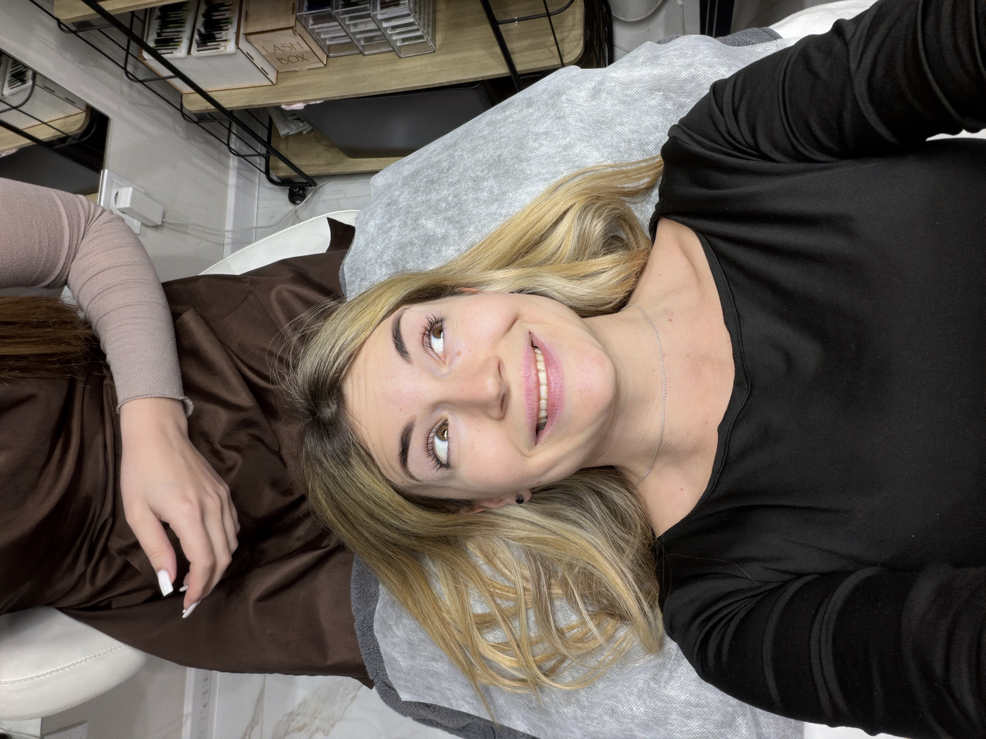 A young woman with blond hair is lying on a treatment table, looking up and smiling. She is wearing a black long-sleeved shirt and a silver necklace. Her hair is spread out on a gray disposable covering on the table. Her eyes are brown and her makeup appears fresh. A manicured hand with white nail polish rests on the brown fabric of the table next to her. Above the table, there is a shelf with various small containers labeled "LASH BOX" and others. The scene appears to be an interior of a beauty salon. The exact location in Budva, Montenegro is not identifiable from the visual information provided.
