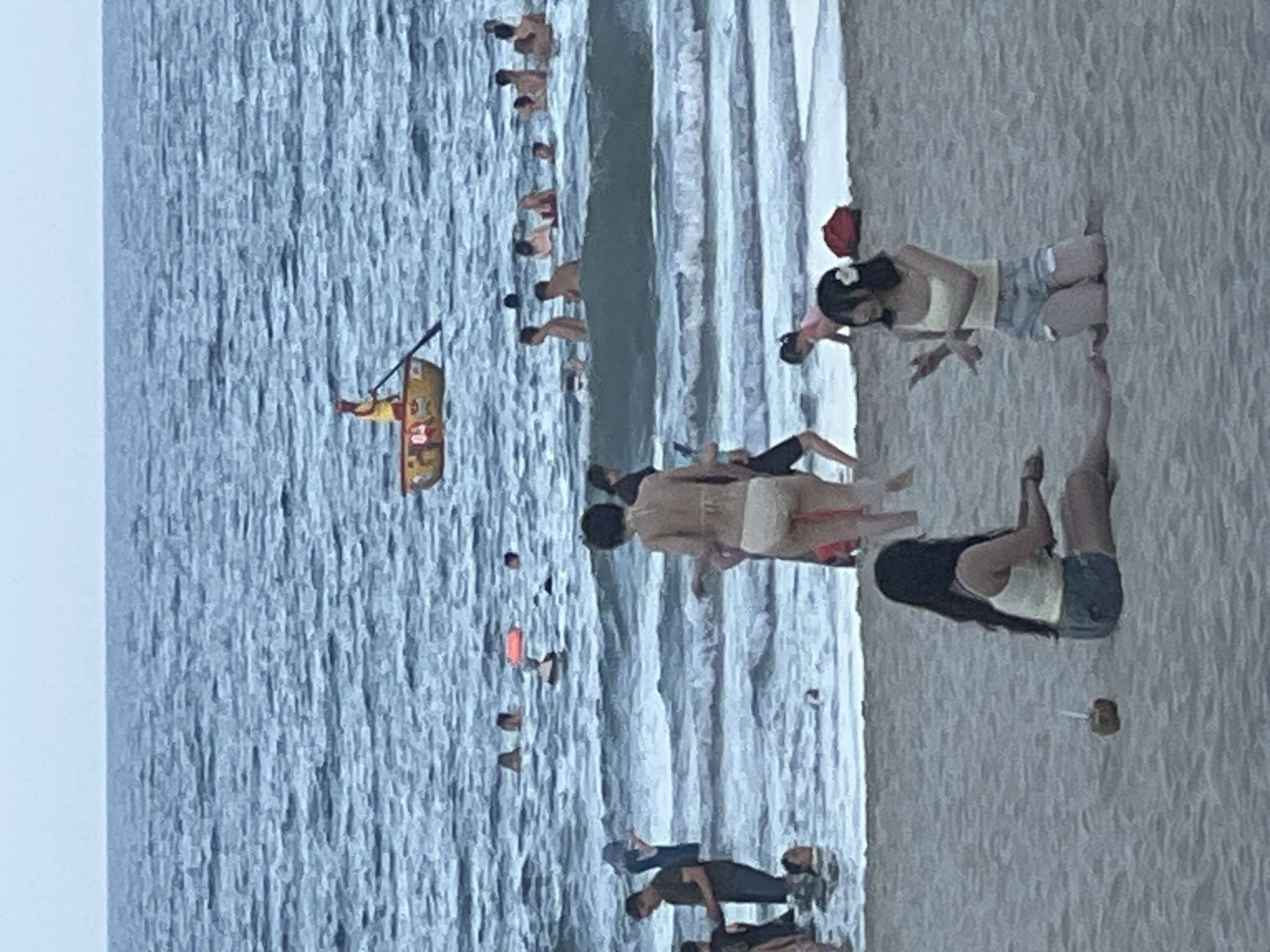 The image depicts a crowded beach scene in Đà Nẵng, Vietnam, likely during daylight hours given the brightness. The foreground is dominated by sandy beach where several people are relaxing. One person is lying down, facing away from the camera, wearing a black tank top and denim shorts. Another person, also lying down, is wearing a dark green shirt and dark pants. Further into the beach, a woman with dark hair adorned with a white flower is sitting, wearing a yellow tank top and denim shorts, with her legs crossed. Next to her, another individual in a pink top is partially visible.
The middle ground transitions to the ocean, with gentle waves lapping at the shore. Several people are wading or swimming in the water. A group of individuals are visible in a line, standing in the shallow water. Further out, a person in a yellow kayak is being paddled by someone else. Scattered throughout the water are other individuals enjoying the sea.
The background is filled with the expanse of the ocean, with more people visible in the distance, appearing as small dots against the water. The sea appears somewhat choppy, with distinct wave patterns. The overall atmosphere seems relaxed and summery, with a clear indication of a popular tourist destination. No text is visible in the image.