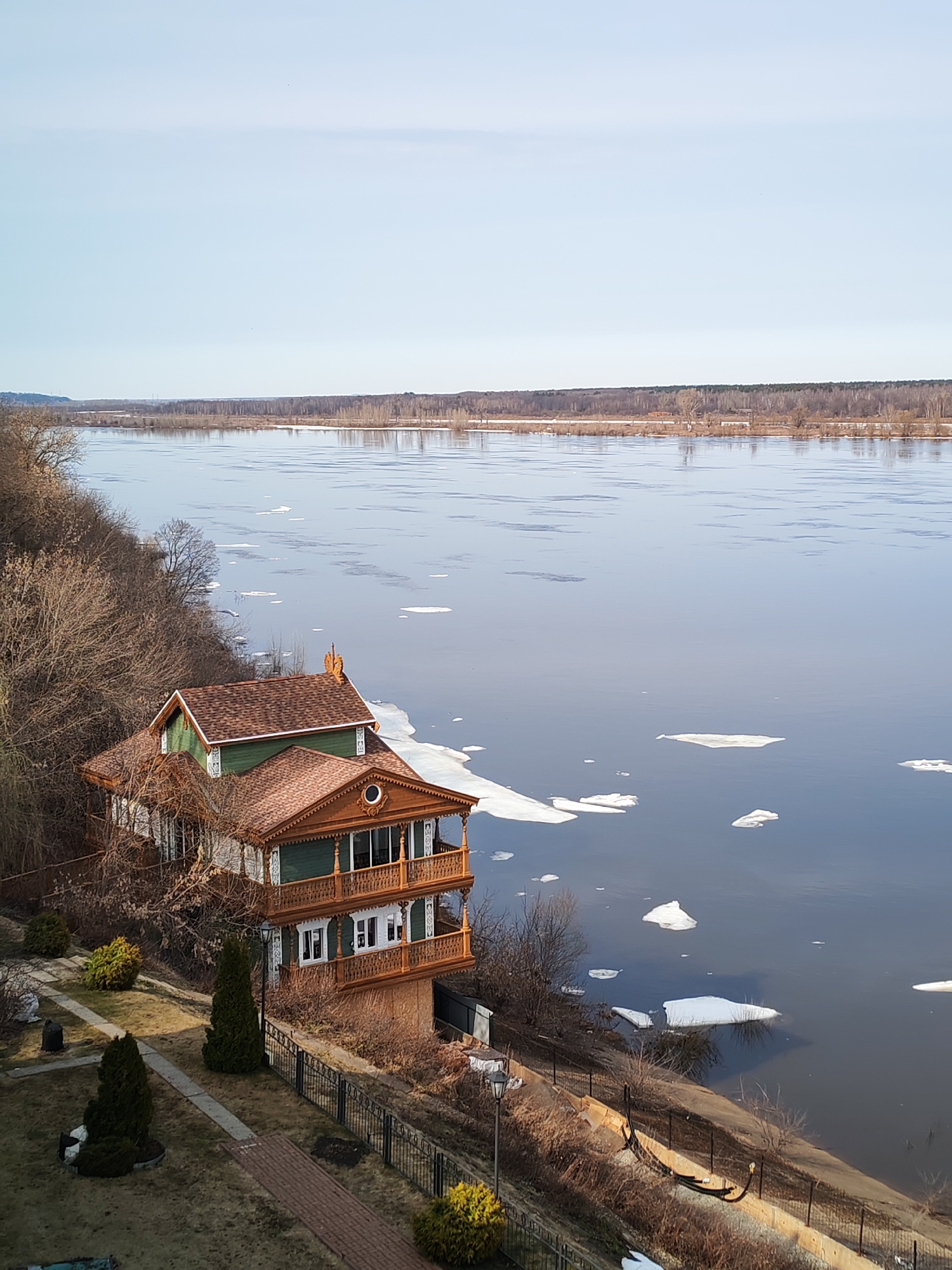 The image shows a two-story wooden house situated on a slope overlooking the Kama River in Sarapul, Russia. The river is partially covered with ice floes. The house is surrounded by trees and a landscaped yard with bushes and a paved path. A metal fence runs along the edge of the slope. A row of trees is visible on the opposite bank of the river. The sky is light blue.