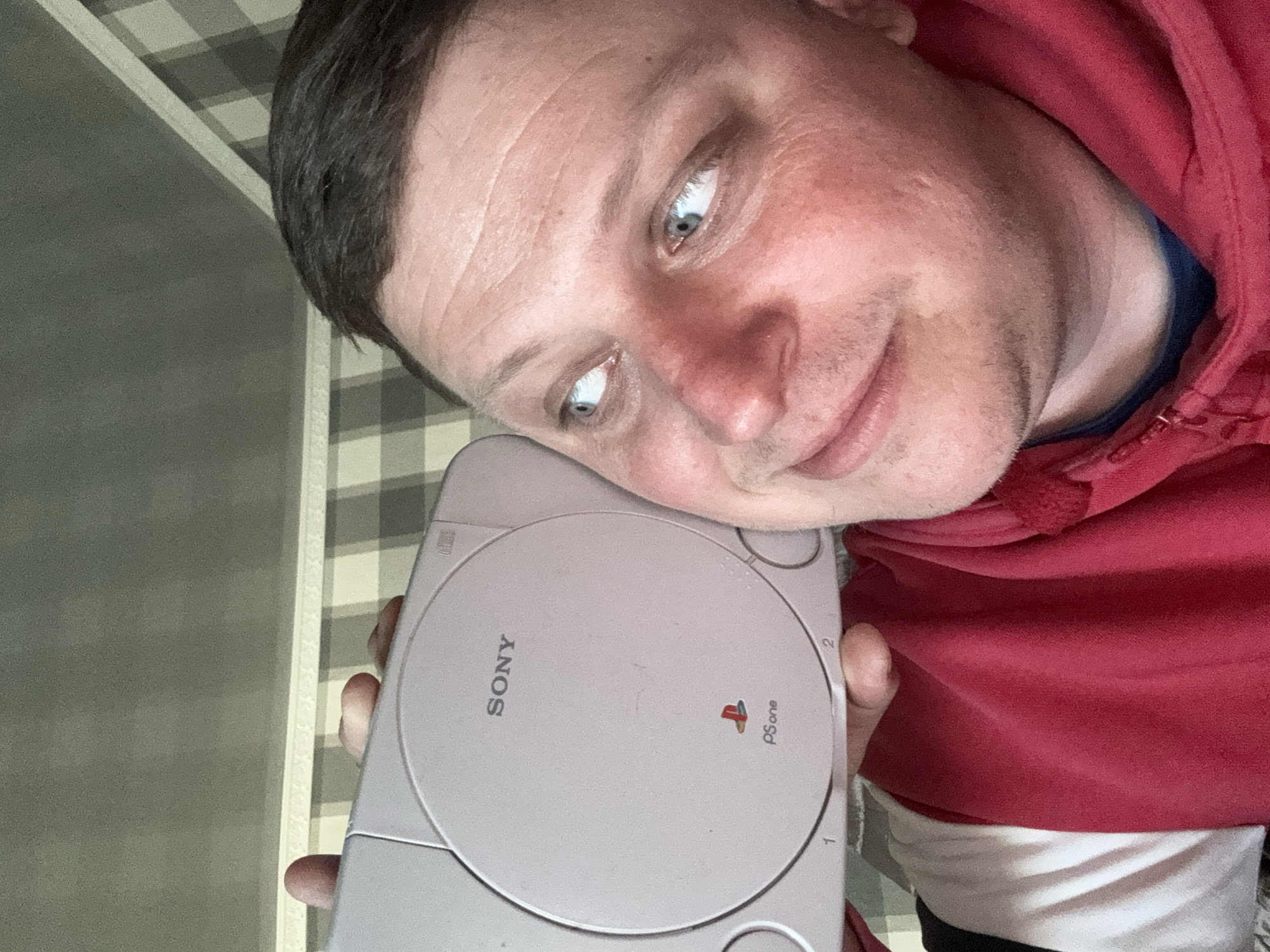 A close-up, slightly rotated selfie shows a man with fair skin and short, dark hair. He has light blue eyes and a slightly smirking expression. He is wearing a red hoodie with a blue undershirt visible at the neckline. The man is holding a light gray Sony PSone (PlayStation One) video game console. The console is facing the viewer, displaying the "Sony" and "PSone" logos, as well as controller ports labeled "1" and "2".
The background appears to be a wall with a checkered pattern. The location is unknown.