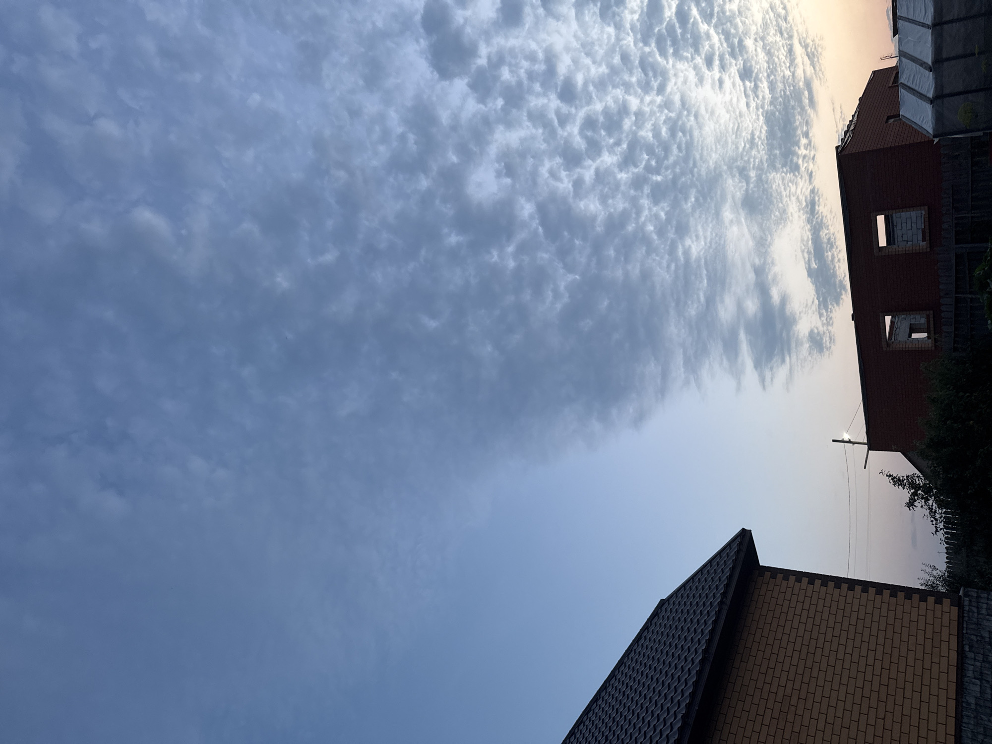 The image shows a partially constructed building and a finished house with a dark tiled roof. The sky is predominantly blue with a band of patchy, textured white clouds stretching across the upper right portion of the frame. The lighting suggests it is either early morning or late evening, with a subtle warm glow on the horizon. In the foreground, the side of a yellow brick house is visible, with a dark roofline. To the right, the brick structure of an unfinished building with open window frames is present, as well as a greenhouse-like structure with a white tarp. A wooden fence runs along the bottom of the image. Power lines and a utility pole are visible in the middle distance. The scene appears to be in a semi-rural or suburban area, consistent with a village or settlement in Russia.