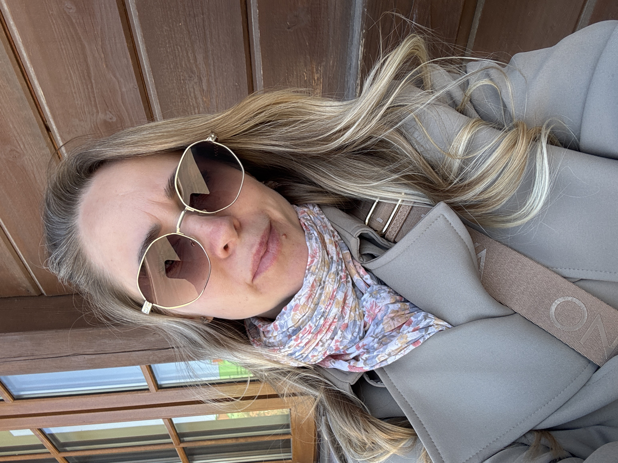 A woman is posing for a photograph. She is wearing sunglasses, a patterned scarf, and a beige trench coat with a branded shoulder strap. Her hair is blonde with darker roots. The background consists of wooden planks and a window with a multi-pane design. The location is Chaikovsky, Russia.