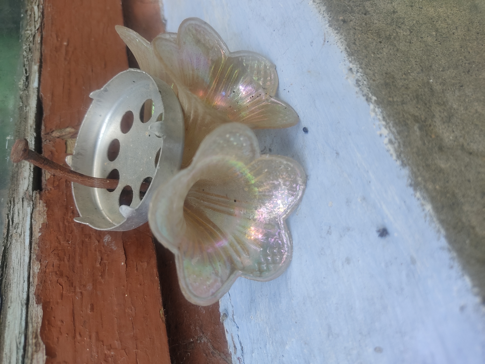 This media file shows a close-up of two iridescent, bell-shaped glass ornaments attached to a rusted nail. The ornaments appear to be decorative, possibly for a garden or outdoor decoration, given the rustic setting. They are mounted on what looks like weathered wooden siding, painted a reddish-brown, and against a light blue painted surface. A rusty nail protrudes from the side of the ornaments and is embedded in the wood. The glass has a pearlescent quality, reflecting light and showing subtle hues of pink and green. The background is somewhat blurred, but suggests an outdoor environment with possibly a textured, gray surface visible on the right. The lighting appears to be natural, suggesting daytime. The overall impression is one of a slightly weathered, perhaps forgotten, decorative element in a rustic outdoor setting. No people are visible, and there is no text. The location context is Voludryntsi, Ukraine.