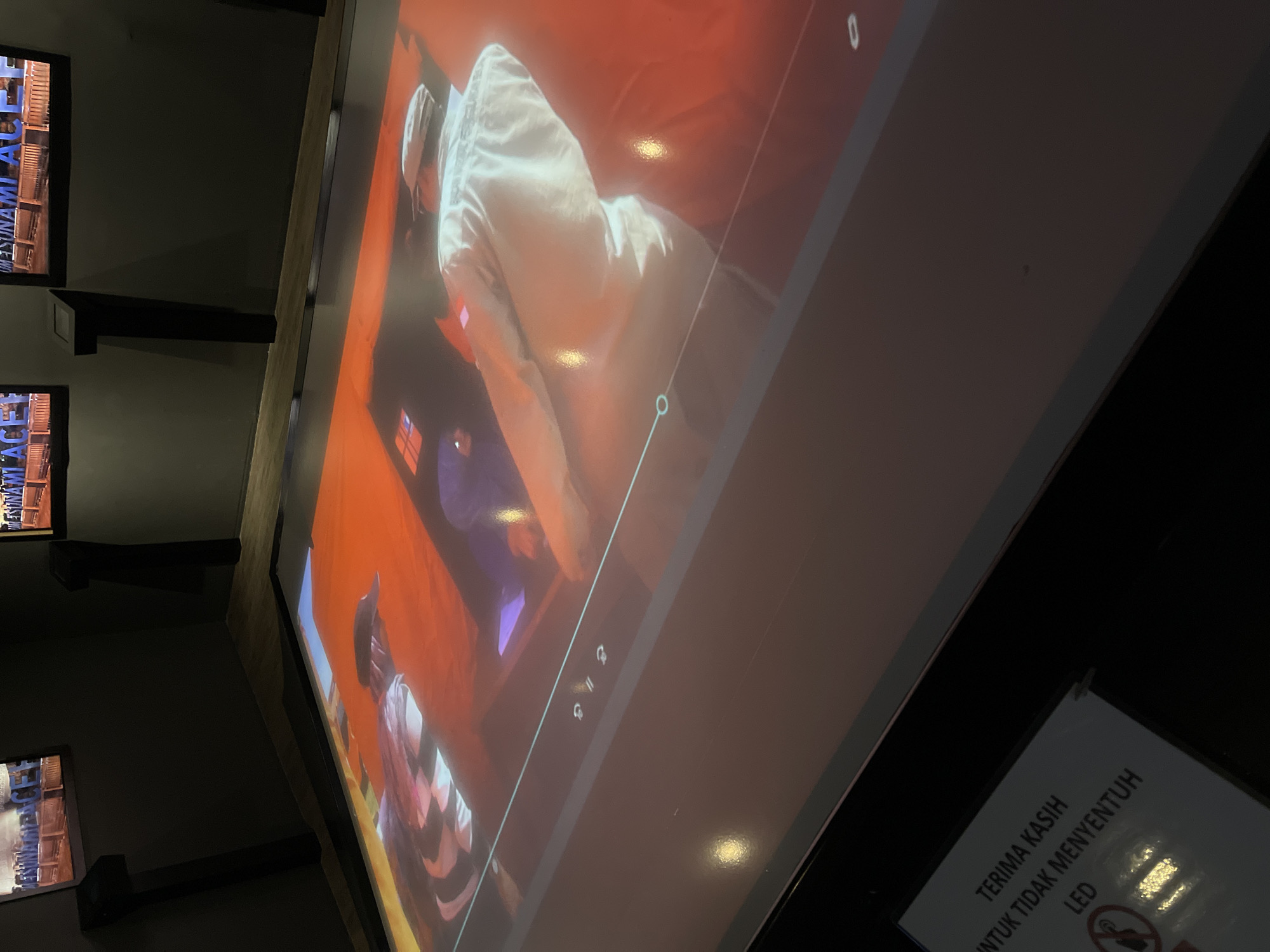 The image depicts an indoor exhibition space, likely a museum or educational center, in Banda Aceh, Indonesia. A large, interactive screen is projected onto the floor, displaying a video with two individuals. One person, wearing a cap and a checkered shirt, is visible in the upper left portion of the screen. The other person, wearing a white jacket and a cap, is more prominent in the center and right of the screen. The video appears to be a historical or documentary piece, as suggested by the text on the screens in the background which reads "TSUNAMI ACEH". A sign on the lower left of the image, written in Indonesian, says "TERIMA KASIH UNTUK TIDAK MENYENTUH LED" which translates to "Thank you for not touching the LED". The environment is dimly lit, with the screens serving as the primary light sources. The overall impression is of an exhibit designed to inform visitors about the impact of the 2004 Indian Ocean tsunami in Aceh.