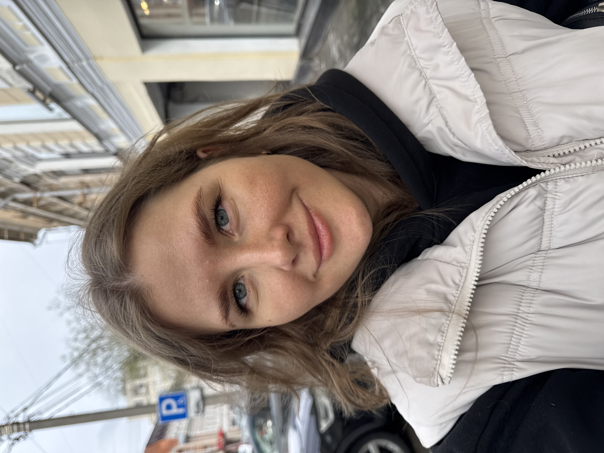 A woman with light brown hair, wearing a light-colored puffer jacket over a black hooded sweatshirt, is facing the camera with a slight smile. Her eyes are light blue. The background appears to be an urban street scene. In the background, there's a "P" parking sign, architectural details of buildings, and parts of a car. Considering the signage, architecture and the background details, the location is plausibly in Perm, Russia.