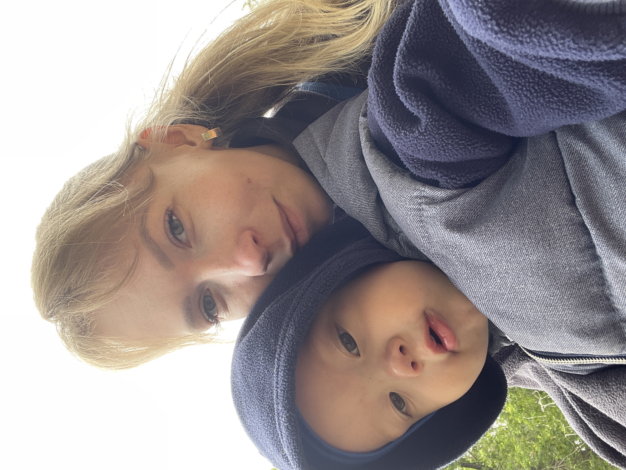 A close-up, eye-level outdoor selfie captures a woman and a baby. The woman, positioned in the upper right, has fair skin, light-colored eyes, a neutral expression, and long blonde hair with some strands appearing windblown. She wears a small, gold-colored, rectangular hoop earring in her visible right ear and a dark-colored garment, possibly a jacket or fleece, with a blue accent visible on the collar area. The baby, situated in the lower left, has dark eyes, a small nose, and slightly parted lips. The baby is wearing a dark blue fleece hooded top, with the hood up, revealing a lighter blue lining inside. The baby is secured in a grey, denim-textured baby carrier that features visible stitching and a zipper on the front. The background consists primarily of a bright, overcast sky. Green foliage is visible in the lower left portion of the frame, suggesting a natural outdoor environment. Both individuals are looking directly into the camera.