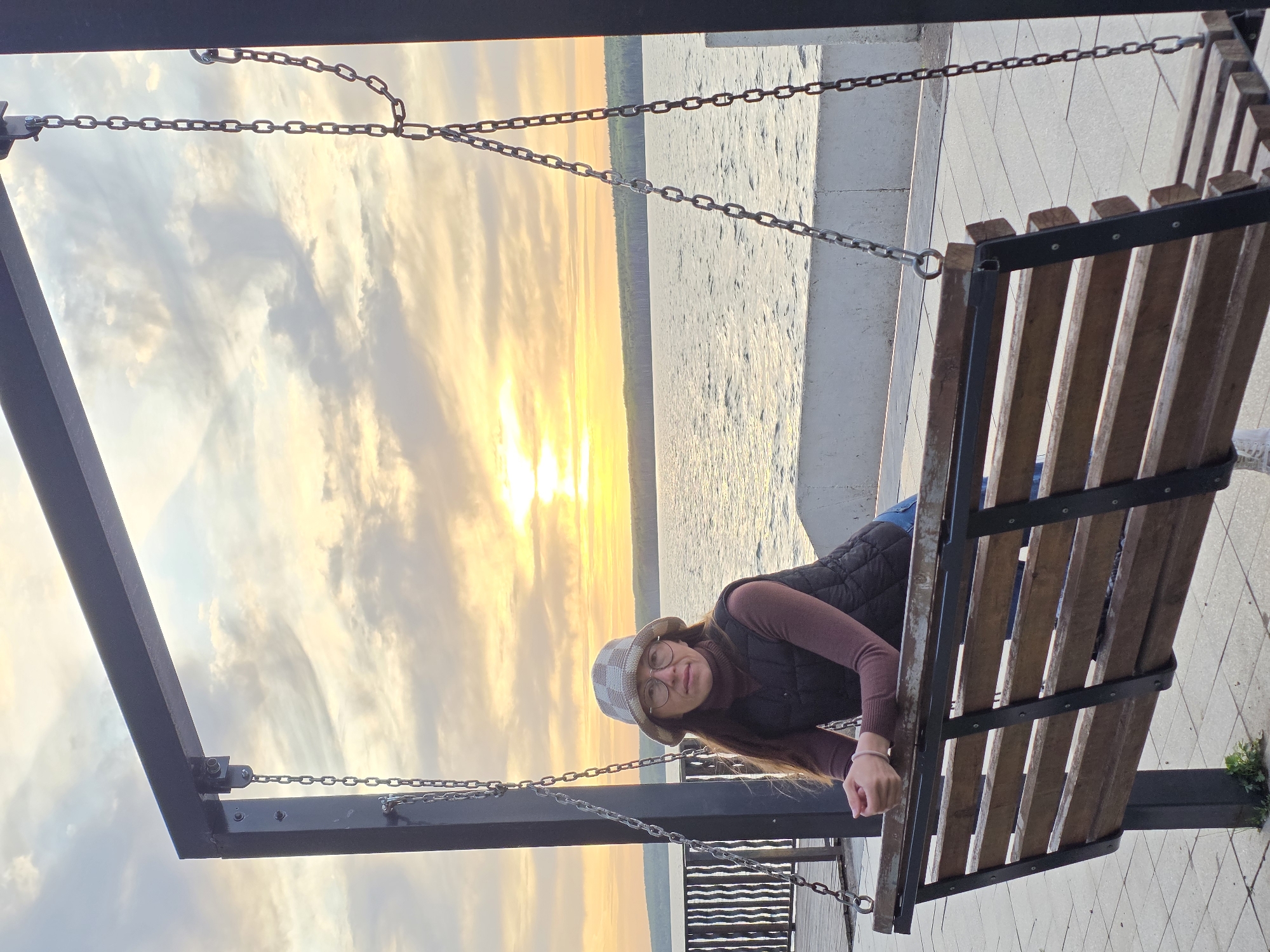 A photograph taken in Chaikovsky, Russia, depicts a woman with long brown hair, wearing glasses, a brown turtleneck, a black vest, and a checkered hat. She is leaning on a swing bench made of wood and metal. The swing is suspended by chains. The background shows a sunset over water. The ground near the swing is paved with light-colored tiles.