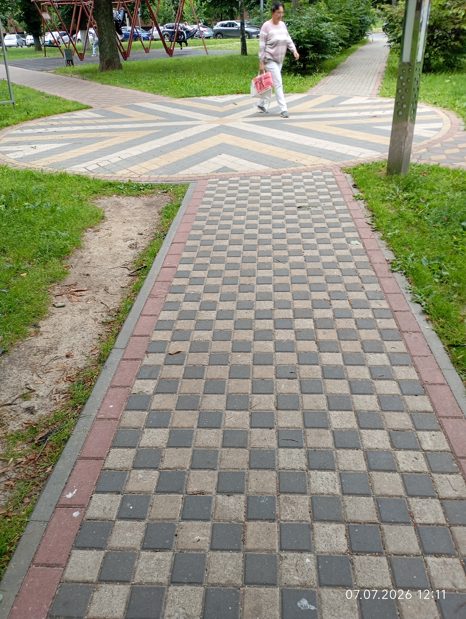 The image depicts a park path in Homyel, Belarus, featuring a woman walking across a decorative pavement area. The path is paved with interlocking stones in a checkered pattern of grey and beige, bordered by red and grey stones. To the left of the path is a strip of dry soil and sparse grass, and further left, a playground with swings and metal structures. To the right, a metal pole with circular perforations stands next to the path, with a grassy area beyond. The woman, wearing a striped sweater and white pants, carries two shopping bags, one red and one white, as she walks away from the viewer towards the playground. In the background, trees and parked cars are visible. A timestamp in the lower right corner reads "07.07.2026 12:11".