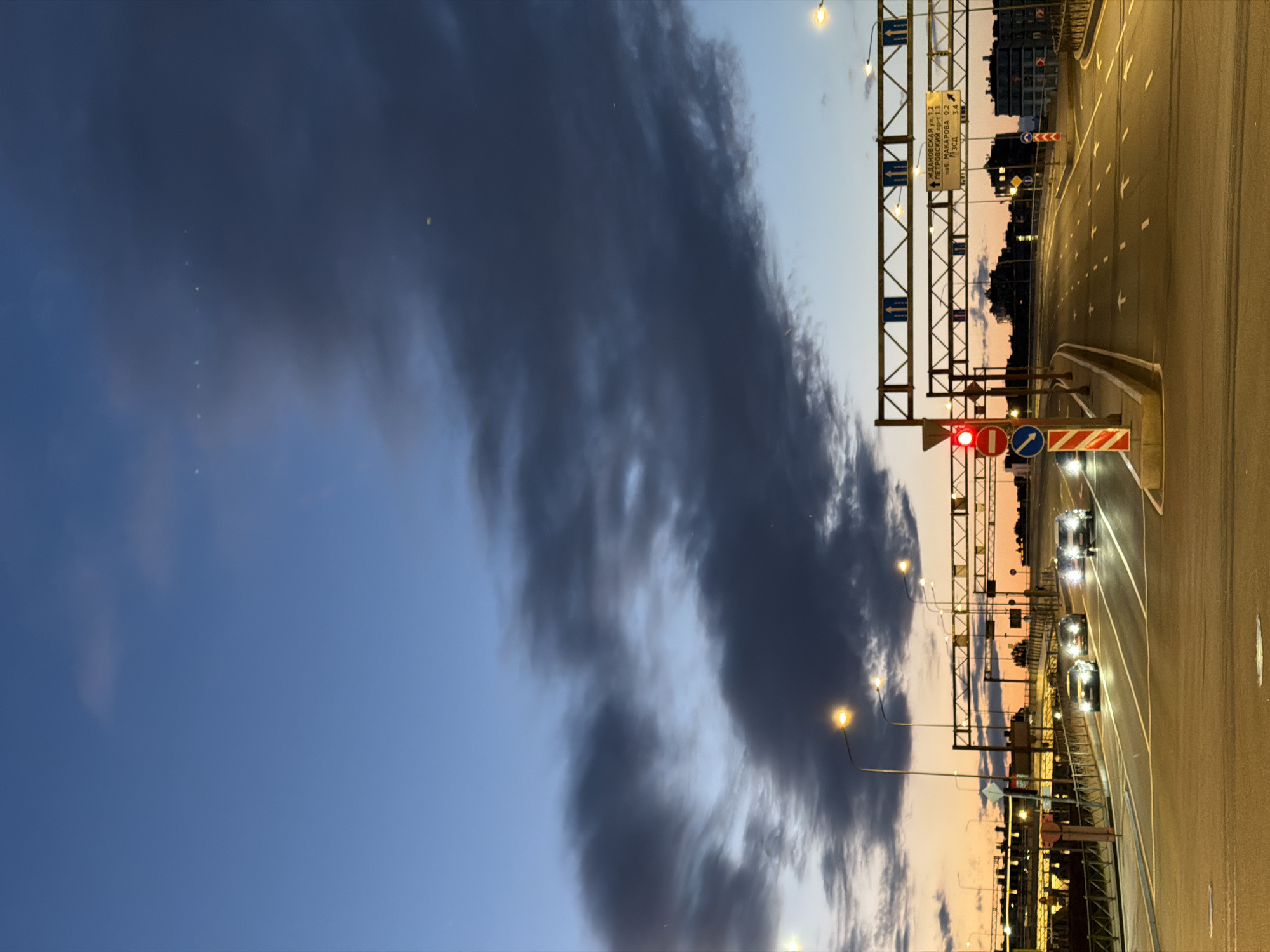 The image depicts a highway overpass in Saint Petersburg, Russia, at dusk. Several cars are driving on the multi-lane road, their headlights illuminated. Overhead gantries display directional signs, with some indicating routes to Zhdanovskaya Street, Petrovsky Prospect, and Makarova Embankment. A traffic light shows red, and various road signs are visible, including circular blue signs with white arrows and a red and white striped barrier. The sky is a gradient of deep blue and orange, with dark clouds obscuring parts of the horizon. Streetlights are on, illuminating the road and surrounding infrastructure, which includes buildings in the distance and a metal fence along the side of the overpass.
