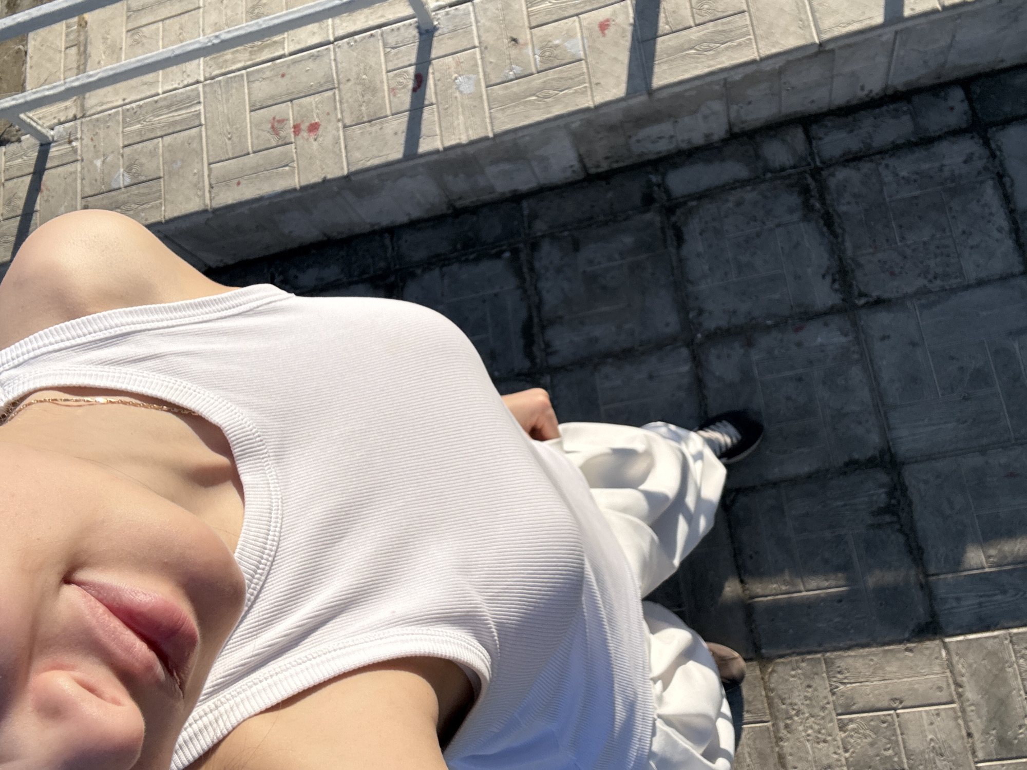 The image presents a close-up, elevated perspective of a person with fair skin. The individual is wearing a ribbed white tank top and white pants. A thin gold necklace is visible on their neck. The person's face is partially visible, showing their lips and nose. The background is a paved area with a metal railing along one edge. The location is possibly in an urban environment. The city of Agryz, Russia, cannot be confirmed.