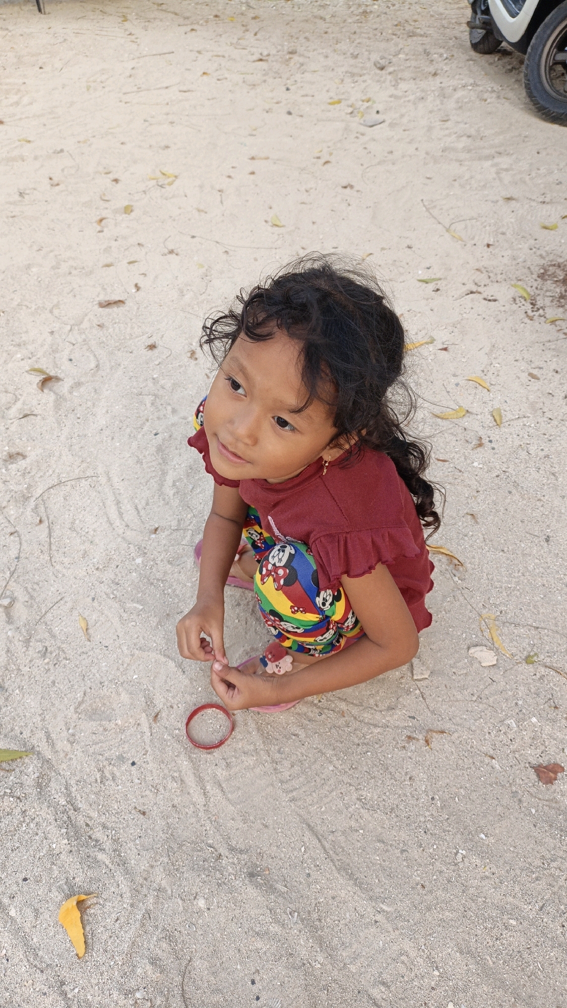 A young girl with dark, curly hair is sitting on a sandy surface, which appears to be a beach or a sandy path, possibly in Cikal, Indonesia. She is wearing a maroon short-sleeved shirt with ruffled edges and colorful shorts adorned with cartoon characters, including Mickey Mouse. Her legs are crossed, and she is looking to her left, with her hands positioned in front of her. A red bracelet lies on the sand near her hands. In the background, a portion of a motorcycle is visible on the right side of the image. The sandy ground is scattered with dry leaves, and the lighting suggests it is daytime.