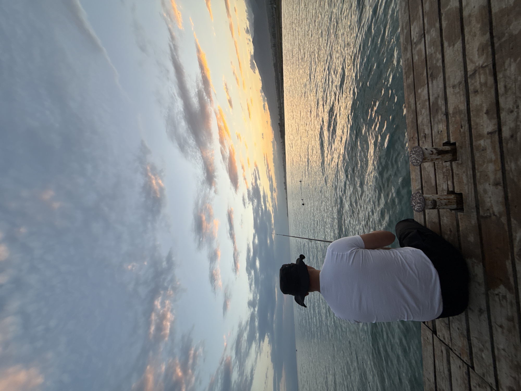A person wearing a white t-shirt and black pants, with a black wide-brimmed hat, sits on a wooden pier in Sary-Oy, Kyrgyzstan, fishing at dusk. The person is facing away from the camera, looking out at a calm body of water, likely Issyk-Kul Lake. The sky is filled with clouds tinted with hues of pink, orange, and blue from the setting sun. Distant mountains are visible on the horizon, silhouetted against the colorful sky. The pier is made of weathered wooden planks, and two metal cleats are attached to it. The water reflects the colors of the sky, creating a shimmering effect. In the distance, a few other people can be seen in the water.