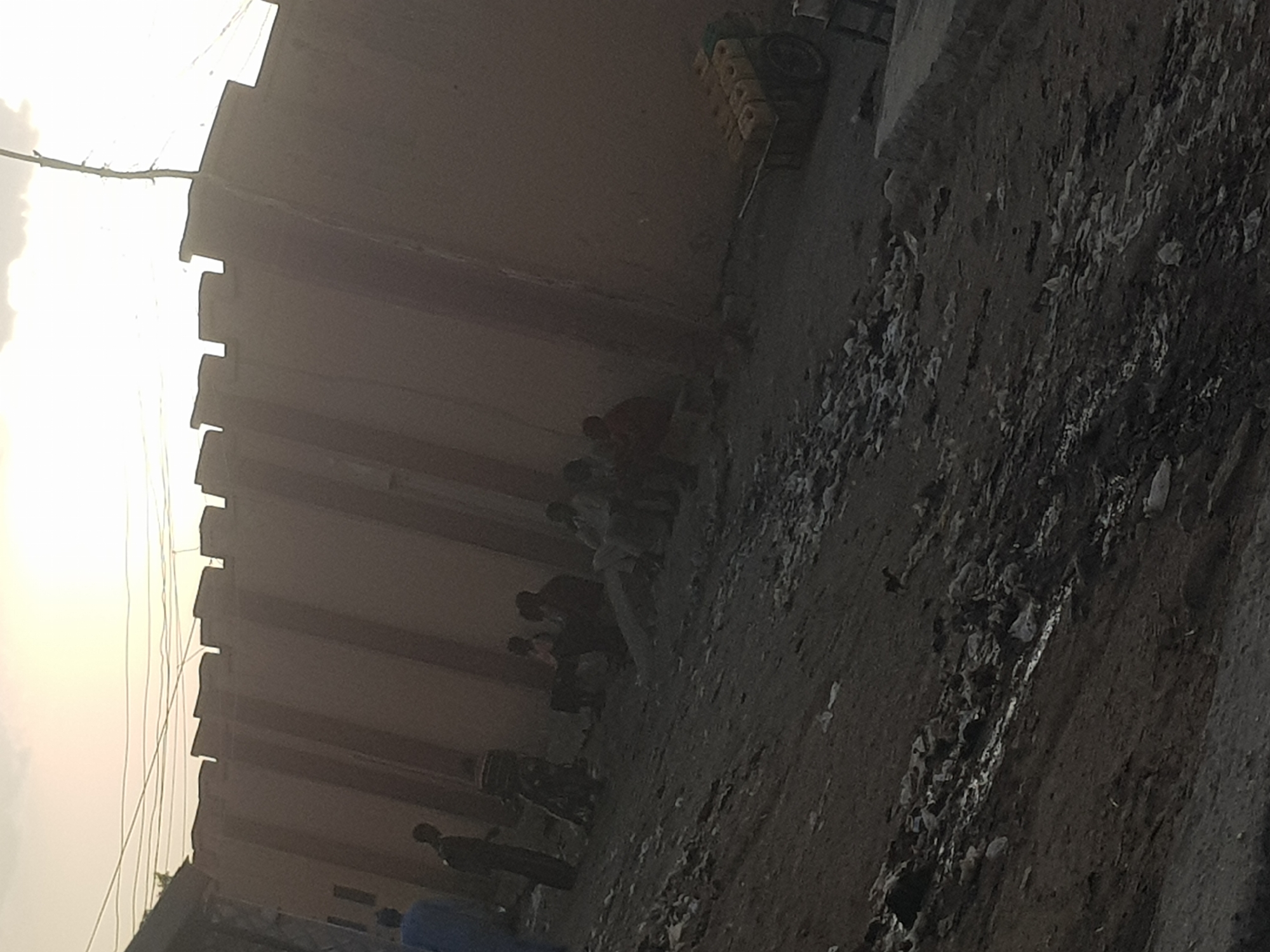 The image depicts a scene in what appears to be a narrow alley in Dutsinma A, Nigeria. A row of people are lined up against a tall, segmented wall. The ground is uneven and covered with debris. In the background, stacked yellow objects are visible near the top of the wall. The lighting suggests it is either early morning or late afternoon.