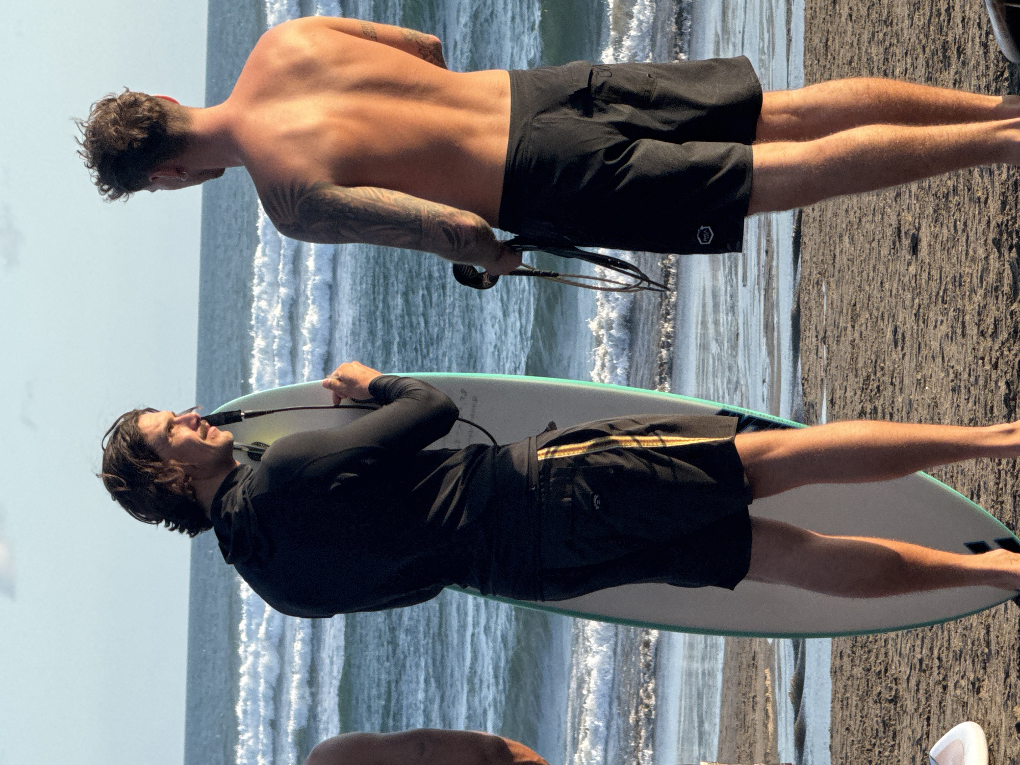 Two fair-skinned men are standing on a black sand beach near the ocean in North Kuta, Indonesia. The man closest to the viewer is lying on a surfboard, wearing a black long-sleeved shirt and black shorts with a tan and black stripe. The other man is shirtless, wearing black shorts, and has tattoos on his left arm and back. He is holding what appears to be a surfboard leash. The ocean waves are visible in the background.