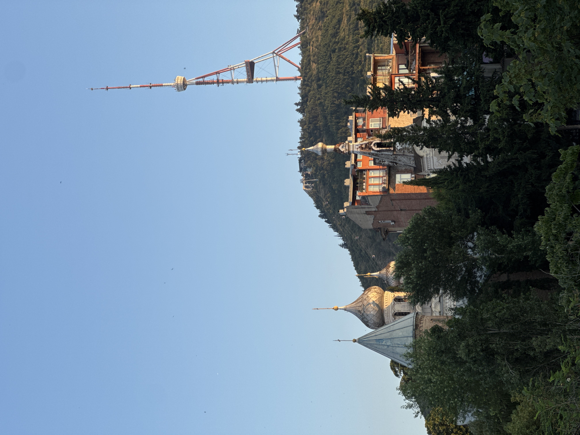The image shows a view of Tbilisi, Georgia, with the Tbilisi TV Tower prominently visible on a forested hill. In the foreground, several domed buildings, likely churches, are partially obscured by lush green trees. Some of these buildings feature distinctive architectural styles with conical or rounded domes. The Tbilisi TV Tower, a tall red and white structure, stands against a clear blue sky. The overall scene depicts a blend of urban architecture, religious buildings, and natural landscape, characteristic of the city's topography.