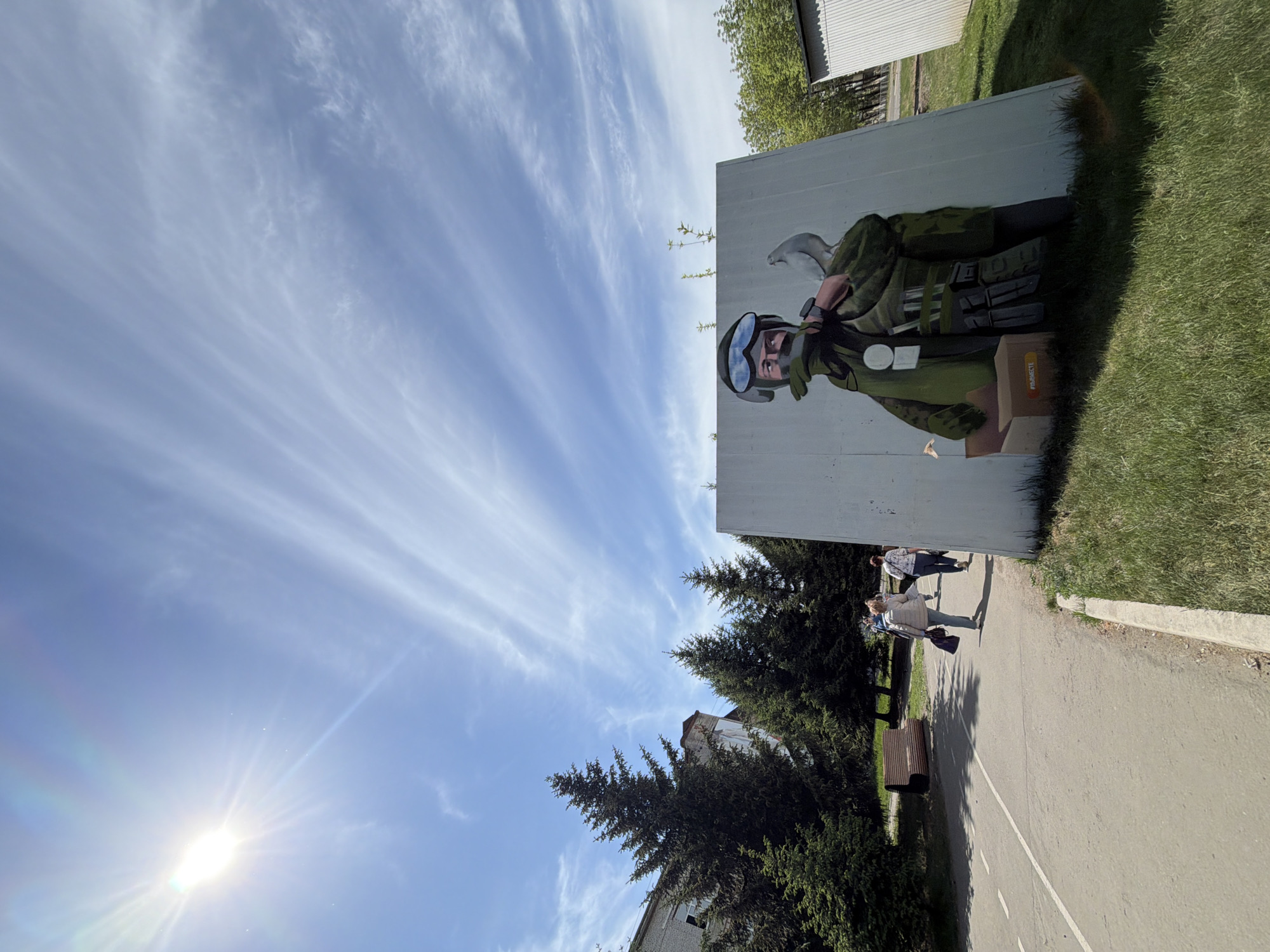 Here's a detailed description of the image:
The image shows an outdoor scene in what appears to be a city park or recreational area. There's a large, light-colored wall or structure with a mural depicting a person in military-style clothing, wearing goggles and holding a bird (possibly a dove) in their hand. The person appears to be emerging from a cardboard box. The mural also contains some text, partially visible.
In the background, there are green trees, grass, and a section of a building. A paved pathway runs along the bottom of the image. Several people are walking along the pathway. A bench is visible near the pathway. The sky is blue with wispy clouds, and the sun is shining brightly. The image seems to be taken on a clear, sunny day.
Based on the tag "#PARRECTE" visible on the cardboard box in the mural, this artwork is located in Birobidzhan, Russia.