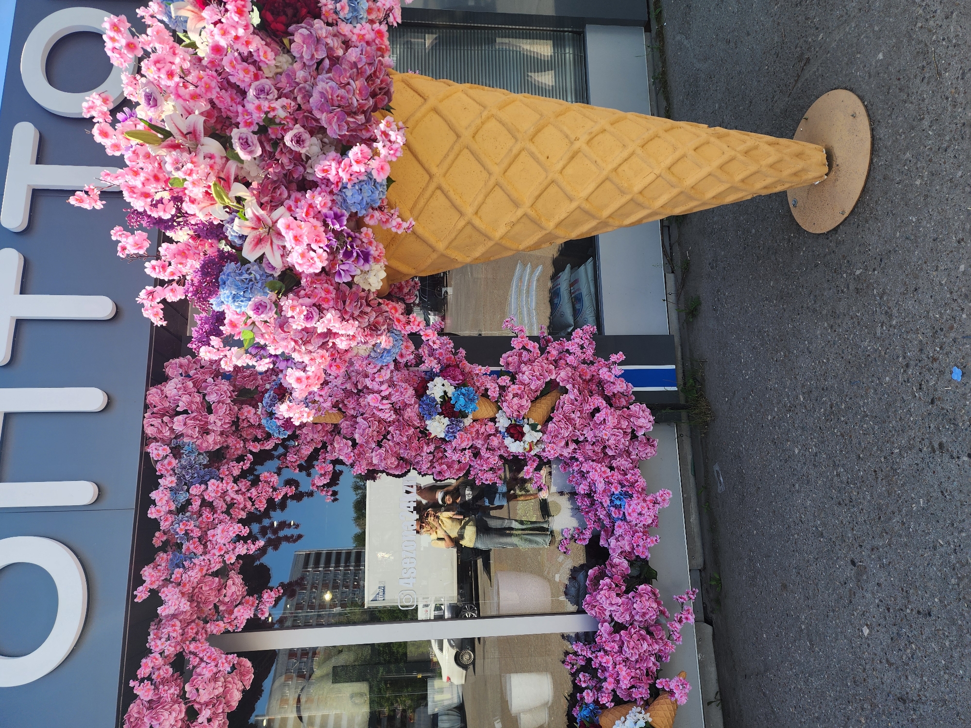 The image shows a storefront decorated with flowers and an ice cream cone sculpture. The store is located in Kazan, Russia, as indicated by the text on the window "КАЗАНЬ". The storefront is light grey, with a white sign reading "OTTO" on the left side. A large, yellow ice cream cone sculpture juts out from the top right of the store, filled with an assortment of pink, purple, and white flowers. Pink flowering branches frame the window on the sides and bottom. The window's reflection shows people, a white car, and nearby buildings. The street pavement is visible on the right side of the image.