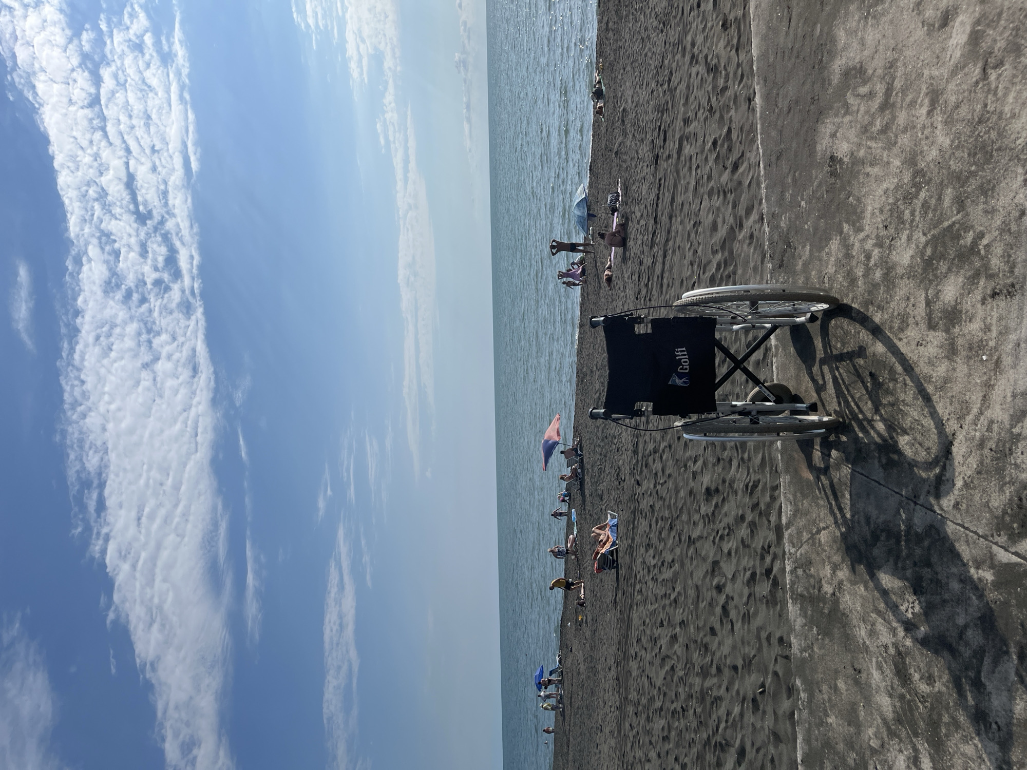 The image depicts a sunny day at Ureki, Georgia, a location known for its magnetic black sand beaches. In the foreground, a wheelchair is positioned on a concrete promenade overlooking the beach. The wheelchair has the brand name "Golfit" visible on its seat. Beyond the promenade, the dark, textured sand of the beach stretches towards the calm, blue sea.
Scattered across the beach are several people enjoying the day. Some are reclining on beach chairs or towels, while others appear to be sunbathing or sitting. A few umbrellas provide shade. The activity suggests a leisurely day at the beach, with individuals relaxing and enjoying the seaside environment.
The sky is a vibrant blue with streaks of white, puffy clouds, indicating fair weather conditions. The sunlight casts distinct shadows, particularly from the wheelchair, suggesting it is likely mid-morning or afternoon. The overall atmosphere appears peaceful and relaxed, typical of a summer day at a popular beach destination. The presence of the wheelchair suggests an effort to make the beach accessible to people with mobility challenges.