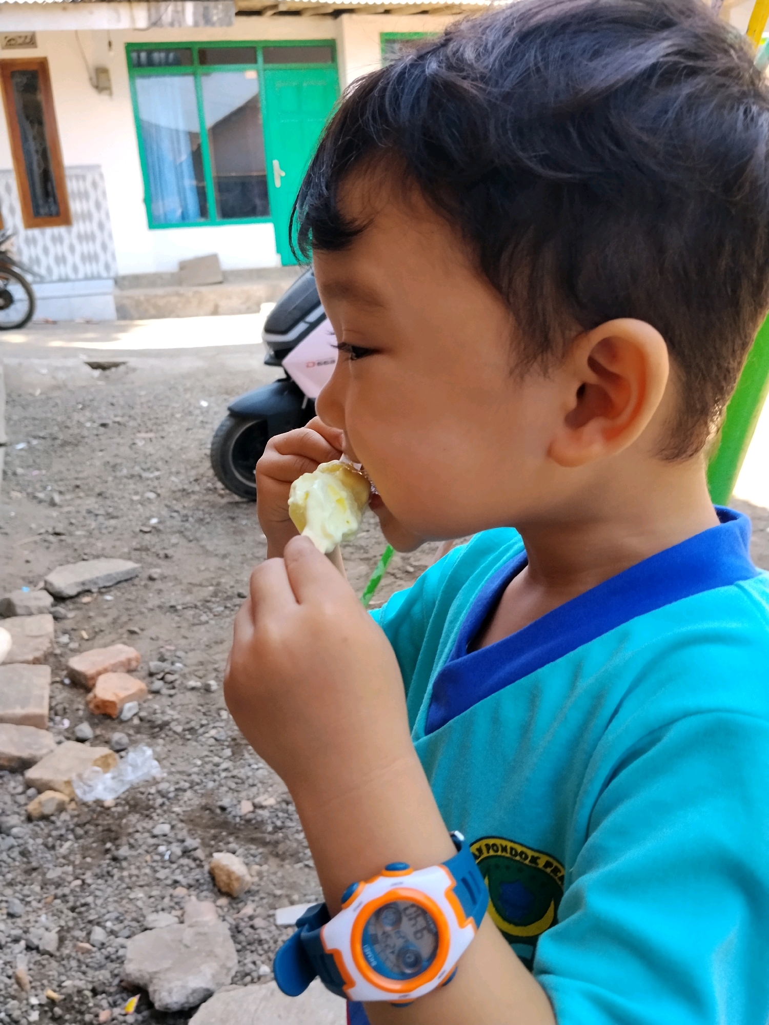 A young boy is captured in a close-up, side-profile shot, from the chest up. He is wearing a teal shirt with a blue collar and has dark hair. His right hand is bringing a piece of light-colored food, possibly a piece of fruit, to his mouth. On his left wrist, he wears a blue, orange, and white digital watch. The background is blurred, but appears to be an outdoor setting with a dirt or gravel ground. There are some scattered bricks and debris on the ground. In the distance, a portion of a white building with green doors and windows is visible. A motorcycle is also partially visible in the background. The overall scene suggests a casual, everyday moment.