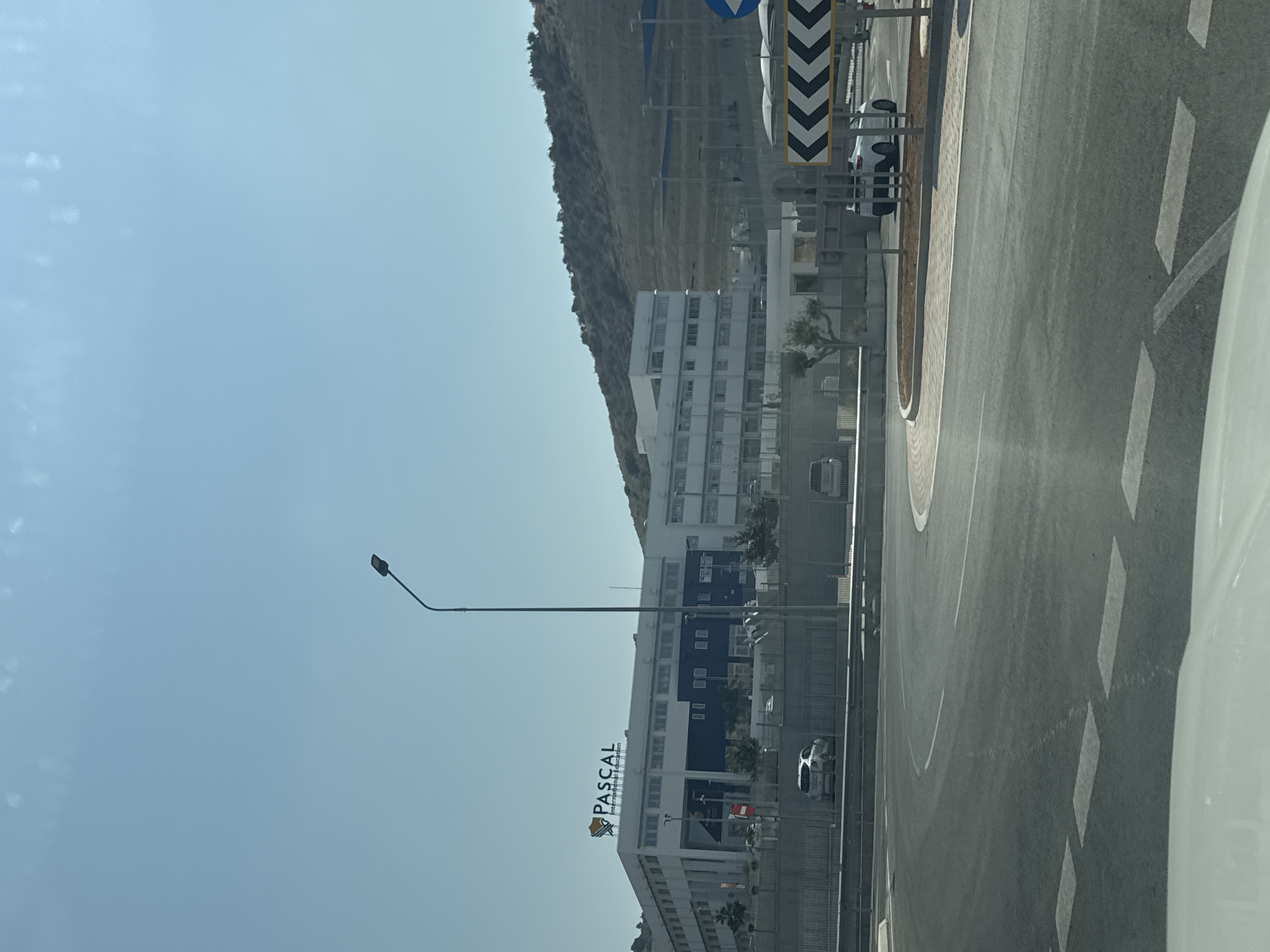 The image shows the Pascal English School in Limassol, Cyprus. It is a large, modern building with a white and blue facade, set against a clear blue sky and a gently sloping, barren hillside. The school's logo, "PASCAL International Education," is prominently displayed on the rooftop. In the foreground, a section of a road with white dashed lane markings is visible, suggesting the viewer is in a vehicle. A street lamp stands tall on the right side of the image. To the right of the school, there are visible structures and a road infrastructure, including directional signs and a white car. The overall impression is of an institutional building located in a semi-arid, sunny environment.