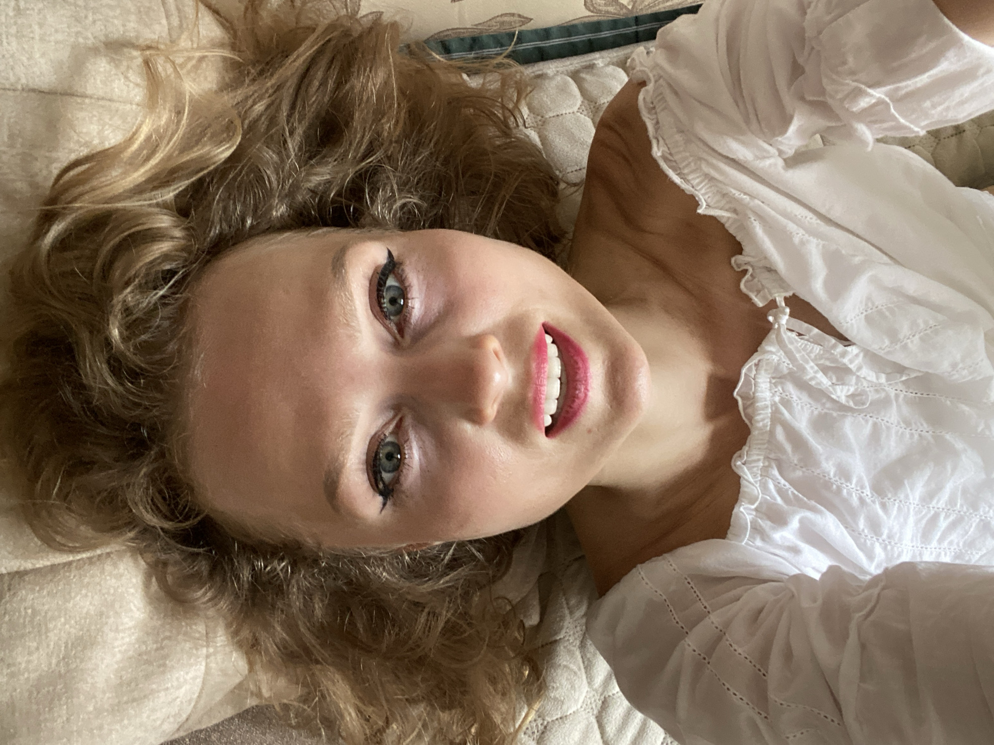 A young woman with blonde, curly hair lies on her back, looking up at the camera. She is wearing a white, off-the-shoulder blouse with ruffled trim and embroidery. Her makeup includes winged eyeliner and bright pink lipstick. Her mouth is slightly open, revealing white teeth. The background appears to be a bed with a textured headboard or pillow. The lighting suggests an indoor setting. There is no information to determine the location in Lida, Belarus.