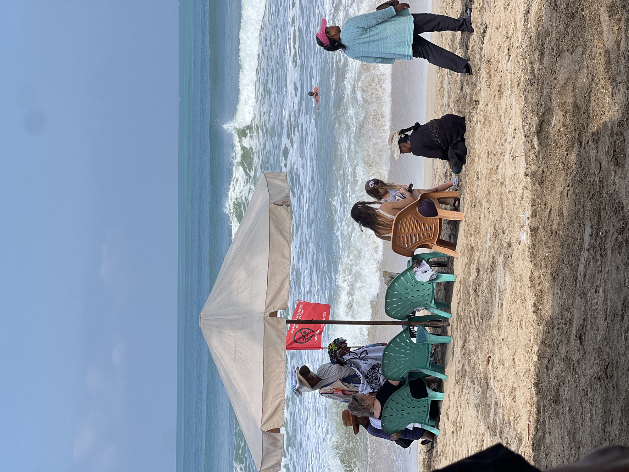 The image captures a beach scene in Kuta, Indonesia, under a clear blue sky. In the foreground, a sandy beach is visible, with several plastic chairs arranged near a large beach umbrella. A group of people are seated on these chairs, some facing the ocean and others interacting with each other. Behind them, the turquoise water of the ocean meets the shore with moderate waves. A person is seen swimming in the water, wearing a bright orange flotation device. Further in the background, the horizon is marked by a faint line of distant land or clouds. A red flag with a prohibition symbol and text in Indonesian and English, indicating "SWIMMING PROHIBITED," is attached to the umbrella pole. People are dressed in casual beach attire, suggesting a relaxed atmosphere despite the warning sign. The overall scene depicts a typical beach day with a mix of leisure and cautionary elements.