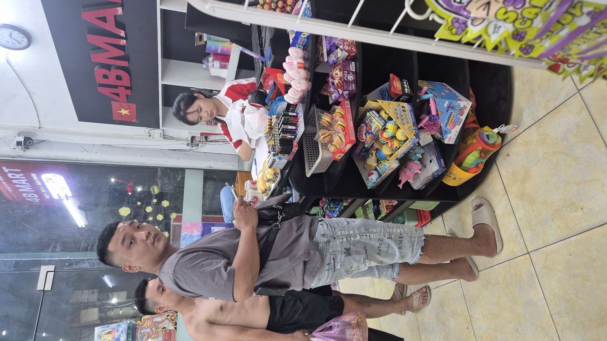 The image shows the interior of a convenience store, likely a "4B Mart" based on the signage. Two men are standing in the foreground, with one man facing the camera, wearing a grey t-shirt and ripped denim shorts, holding a phone and a small dark bag over his shoulder. Another man, shirtless and wearing black shorts, stands to his left, holding a pink plastic bag. A young woman in a red and white shirt is visible behind a counter in the background, interacting with a customer or employee. The counter is laden with various items, including what appear to be candy, lighters, and other small goods. Shelves behind the counter are stocked with more products, including children's toys. The store's floor is tiled with a light yellow color. The overall impression is of a typical small convenience store environment. The presence of "4B MART" signage and the style of the store and products suggests it could be located in Vietnam, specifically Đà Nẵng, given the prompt's context.