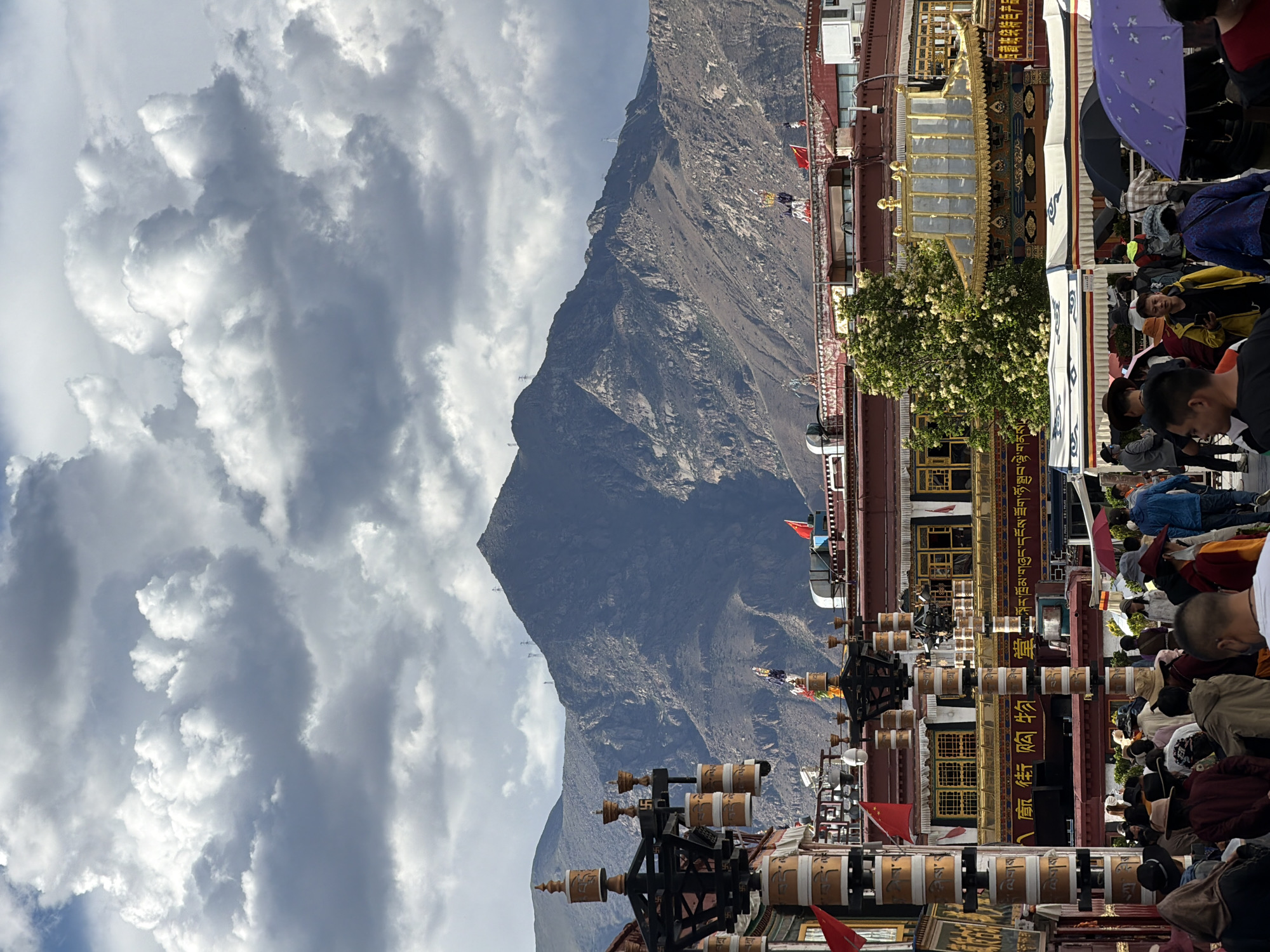 The image, rotated clockwise to its correct orientation, depicts a bustling scene at a traditional architectural complex set against a dramatic mountainous backdrop, likely in China's Chengguan District. In the foreground and midground, ornate buildings feature distinctive golden roofs, intricate carvings, and numerous large prayer wheels adorned with script. Several red flags and colorful prayer flags are visible, alongside a blossoming tree.
Many people are gathered, appearing to be a mix of pilgrims and tourists. They are seen walking, standing, and observing the surroundings; some individuals hold umbrellas (one purple, one black), suggesting either sun protection or a chance of rain. The activity centers around a religious or culturally significant site, indicated by the prayer wheels and temple-like architecture.
The background is dominated by a large, rugged, and somewhat barren mountain, under a sky filled with impressive, textured grey and white clouds. The time of day appears to be daytime, with natural light illuminating the scene.
Visible text includes Tibetan script on the prayer wheels and parts of the buildings. Chinese characters are also visible on signs; for example, "购物" (gòuwù - shopping) can be seen on a horizontal sign, and "寺" (sì - temple/monastery) on a vertical sign, indicating commercial or visitor facilities within the area. The setting suggests a blend of religious devotion, tourism, and daily life in a high-altitude, culturally rich environment.