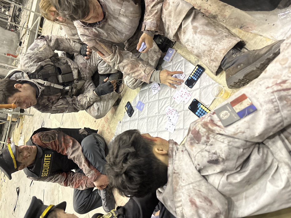 The image depicts an indoor scene in Huangdao District, China, within an environment characterized by exposed concrete flooring, partial wood paneling, and distant scaffolding, indicative of a construction site or derelict structure. Seven individuals are observed seated on the floor around a light grey quilted mat, engaged in what appears to be a card game.
All visible individuals exhibit simulated bloodstains on their attire and faces, consistent with special effects makeup application. In the foreground, a male with short dark hair is shown from the back, wearing a light-colored hooded jacket adorned with a French flag patch and an additional emblem on the right shoulder. To his right, a male in a camouflage tactical uniform with a vest and fingerless gloves reaches for cards on the mat; a small orange cylindrical object is suspended above his head. A female with light hair and simulated facial injuries is partially visible behind him. To the left, a male in a black 'SECURITE' vest, grey pants, and a dark peaked cap with yellow trim observes the scene. Another individual in a similar security uniform is partially visible on the far left. Two additional individuals are partially visible on the right.
The playing surface consists of a quilted mat with several playing cards, some face-up, displaying hearts and spades. Three smartphones, each displaying a calculator interface, are also present on the mat. A clear plastic cup containing an amber liquid is situated near the foreground. A red fire extinguisher is visible in the background, mounted on a wall.