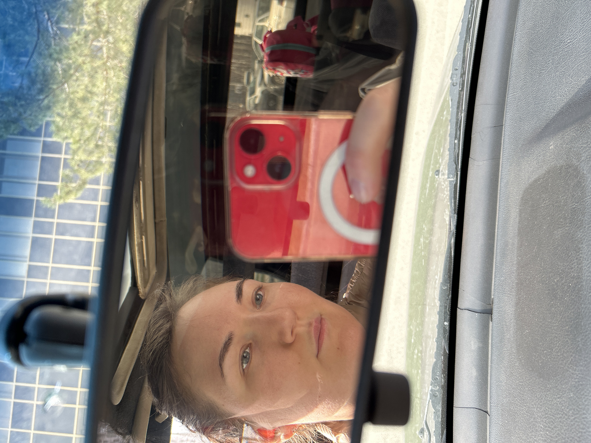A selfie is taken through a vehicle's rearview mirror. The reflection shows a woman holding a red iPhone with a MagSafe charger attached. Outside the vehicle, there's a view of a building with a grid pattern and trees, suggesting a location with modern architecture. The interior of the car is visible around the mirror, including part of the dashboard. There is no way to ascertain whether this image was taken in Chaikovsky, Russia.
