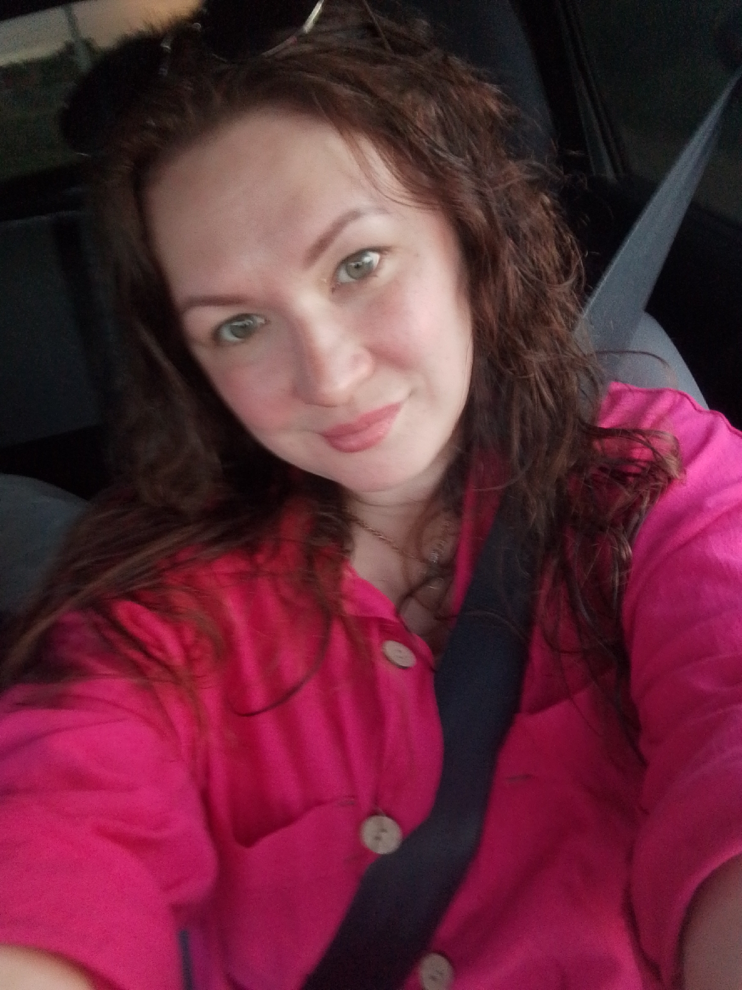 A woman is seated in the driver's seat of a vehicle, as indicated by the presence of a seatbelt. She has fair skin, green eyes, and wavy brown hair. She is wearing a bright pink button-down shirt. The location is Прикамский, Russia. The details are not evident in the image provided.