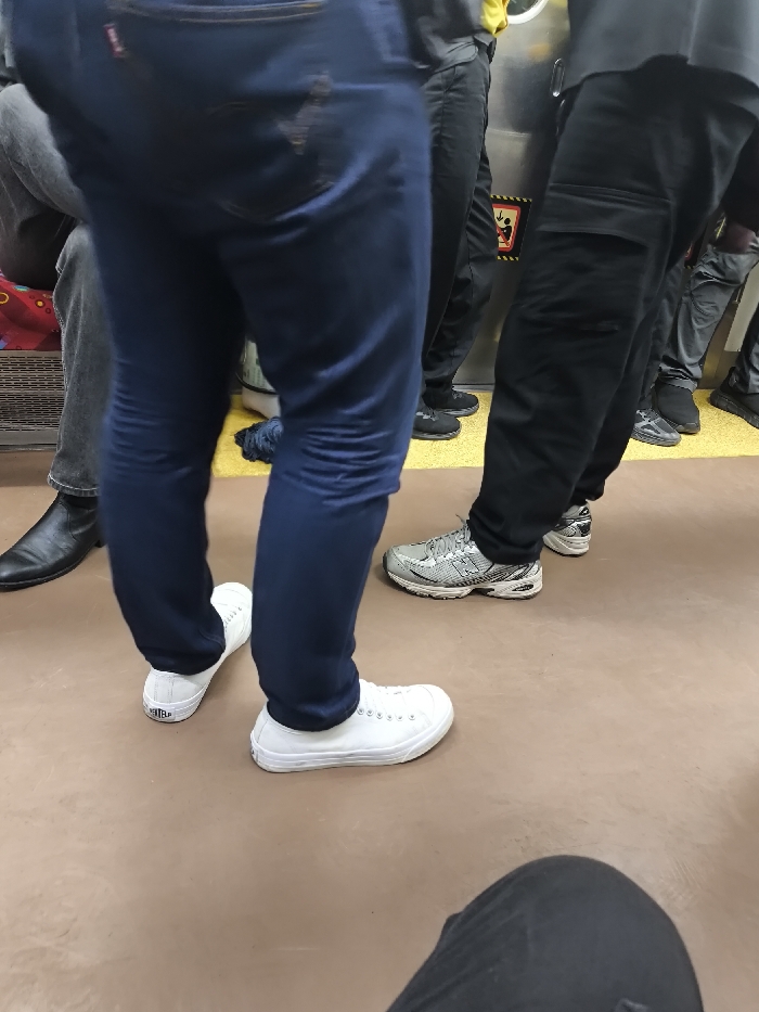 The image presents a ground-level view inside a public transportation vehicle, likely a KRL Commuterline train, situated in Cikarang, Indonesia. The floor in the foreground is a smooth, matte brown-tan, transitioning to a ribbed yellow section further back, adjacent to a metallic interior wall or door.
Multiple individuals are visible from approximately the waist down. In the central foreground, a person stands wearing dark blue jeans with a faded red label on the right rear pocket, and white low-top canvas sneakers with white laces. To their immediate left, a seated individual is visible from the mid-thigh down, dressed in dark grey pants and black pointed-toe dress boots. The seating area features a red and patterned fabric upholstery and a metal grate below.
To the right of the central standing figure, several passengers are positioned. One individual wears black cargo pants and grey and white athletic sneakers. Another, partially obscured, is dressed in black pants and black athletic shoes, with a sliver of a yellow shirt visible above their waist. Further to the right, additional legs in dark pants and shoes are discernible. A small black and red warning sign, featuring a downward arrow and a stylized figure, is affixed to the metallic wall. A crumpled dark blue fabric object rests on the yellow floor section. In the bottom right corner of the frame, the knee and upper thigh of a person wearing black pants are visible, indicating a seated perspective.