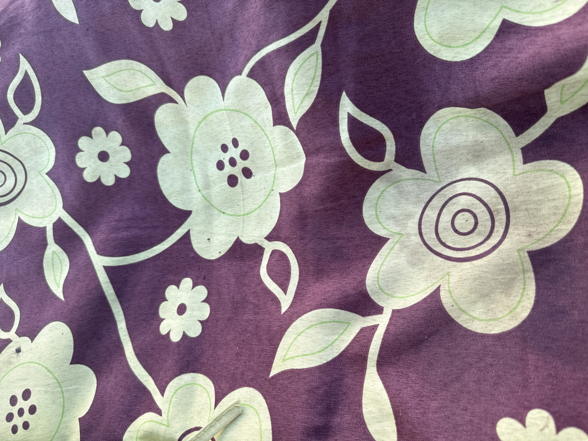 This is a close-up, overhead shot of a purple fabric with a floral pattern. The pattern consists of large, stylized white flowers with green outlines and centers. Some of the flowers have concentric circles in their centers, while others have small dots. There are also smaller, simple white flowers with green outlines scattered throughout the fabric. Green leaves with green outlines are interspersed among the flowers. The fabric appears to be a thin material, possibly cotton or a blend, and it has a slightly wrinkled texture, indicating it might be clothing, bedding, or drapery. The lighting is somewhat harsh, creating shadows that highlight the texture of the fabric. There is no visible text. The scene is static, focusing solely on the textile design.