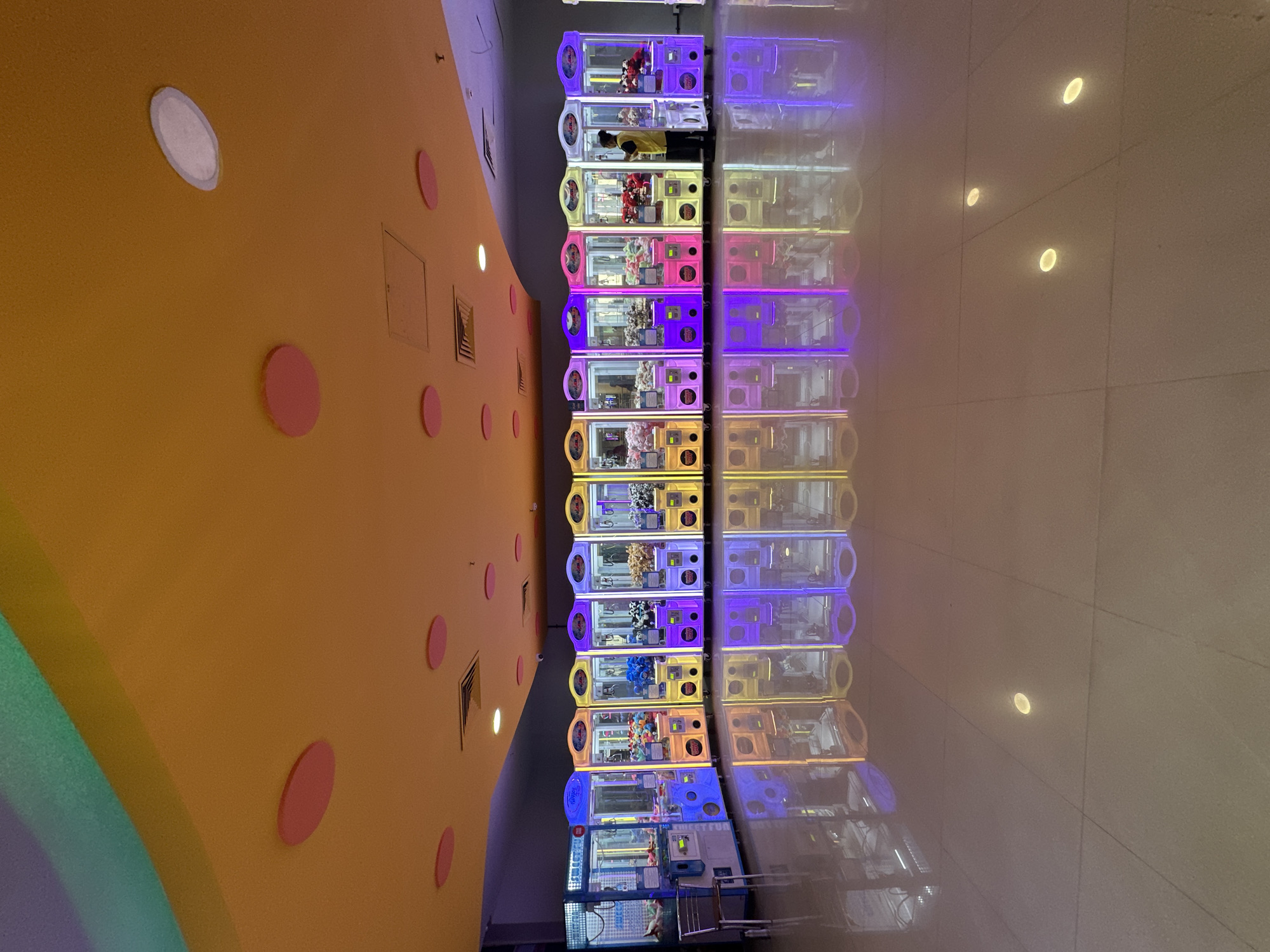 The image depicts a row of claw machines lining a wall in a hallway, likely within a shopping mall or arcade, based on the polished floor and wall design. The machines are colorful, with each seemingly having a different color scheme and filled with various prizes, mostly plush toys. A person, likely an employee, is standing between two of the claw machines. The scene appears to be in Kalyan-Dombivli, India, given the instructions. The wall beside the claw machines is painted yellow and adorned with pink circles. The floor creates a reflection of the claw machines. The lighting is bright, with the machines lit internally. The ceiling has recessed lighting.