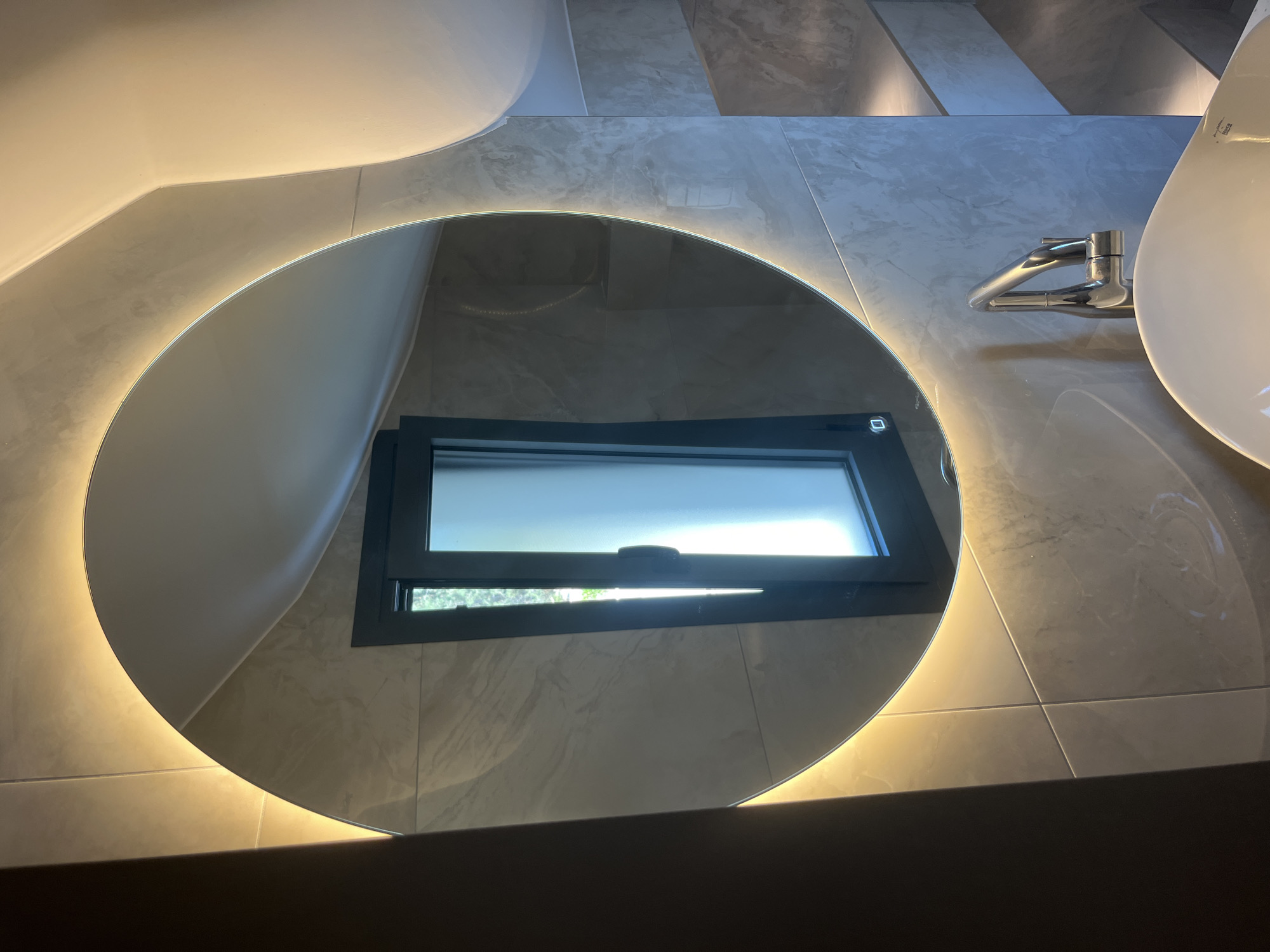 The image depicts a modern bathroom with a large, circular mirror illuminated by a warm LED light around its perimeter. The mirror is mounted on a wall tiled with large, grey marbled-effect tiles. Reflected in the mirror is a dark-framed window, partially open, revealing natural light and greenery outside. Below the mirror, a chrome faucet with a sleek, curved design is visible, positioned over a white ceramic sink. The overall aesthetic is contemporary and minimalist, suggesting a high-end bathroom design. The location is specified as Moni Community, Cyprus.
