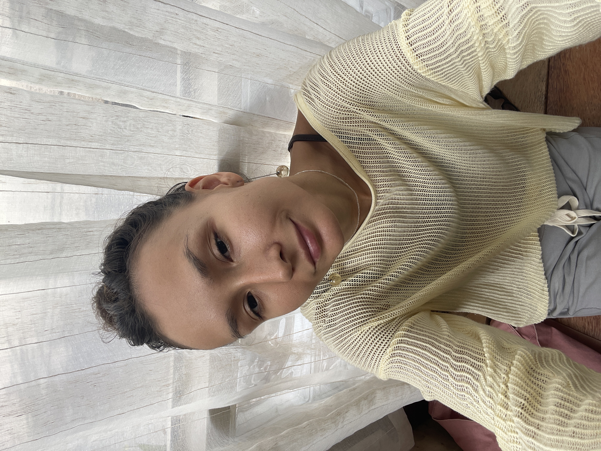 A young woman with dark hair pulled into a bun, wearing a light yellow mesh top and grey pants, is pictured from the chest up, looking directly at the camera. She has subtle makeup on and is wearing a delicate necklace and earrings. Behind her, sheer white curtains are drawn across a window, with glimpses of the outdoors visible through the fabric. The setting appears to be indoors, possibly in Mũi Né, Vietnam, given the light and texture of the curtains, which could be characteristic of the local environment or accommodation.