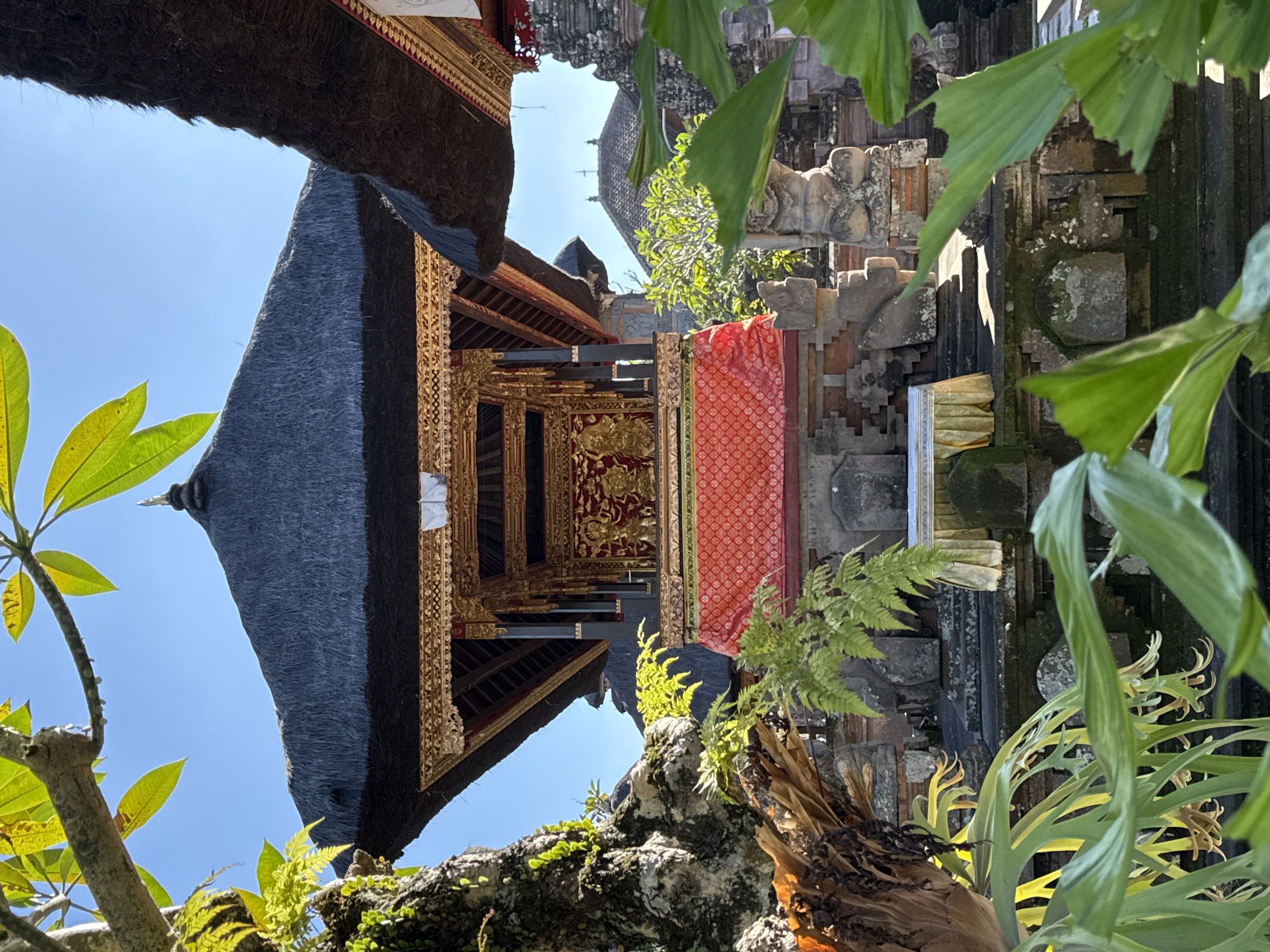 This image captures a detailed view of a traditional Balinese temple structure, likely situated within the lush environment of Ubud District, Indonesia. The central focus is a multi-tiered roofed building with intricate golden ornamentation, covered in dark thatch. A vibrant red and gold fabric drapes across the lower section of this central structure. In front of it, a low table is draped with yellow cloth. The temple is surrounded by stone carvings and moss-covered walls, with lush green foliage framing the scene from the foreground and sides. Sunlight illuminates the scene, casting shadows and highlighting the textures of the natural elements and the temple architecture. The sky is clear blue. No people are visible in the image.