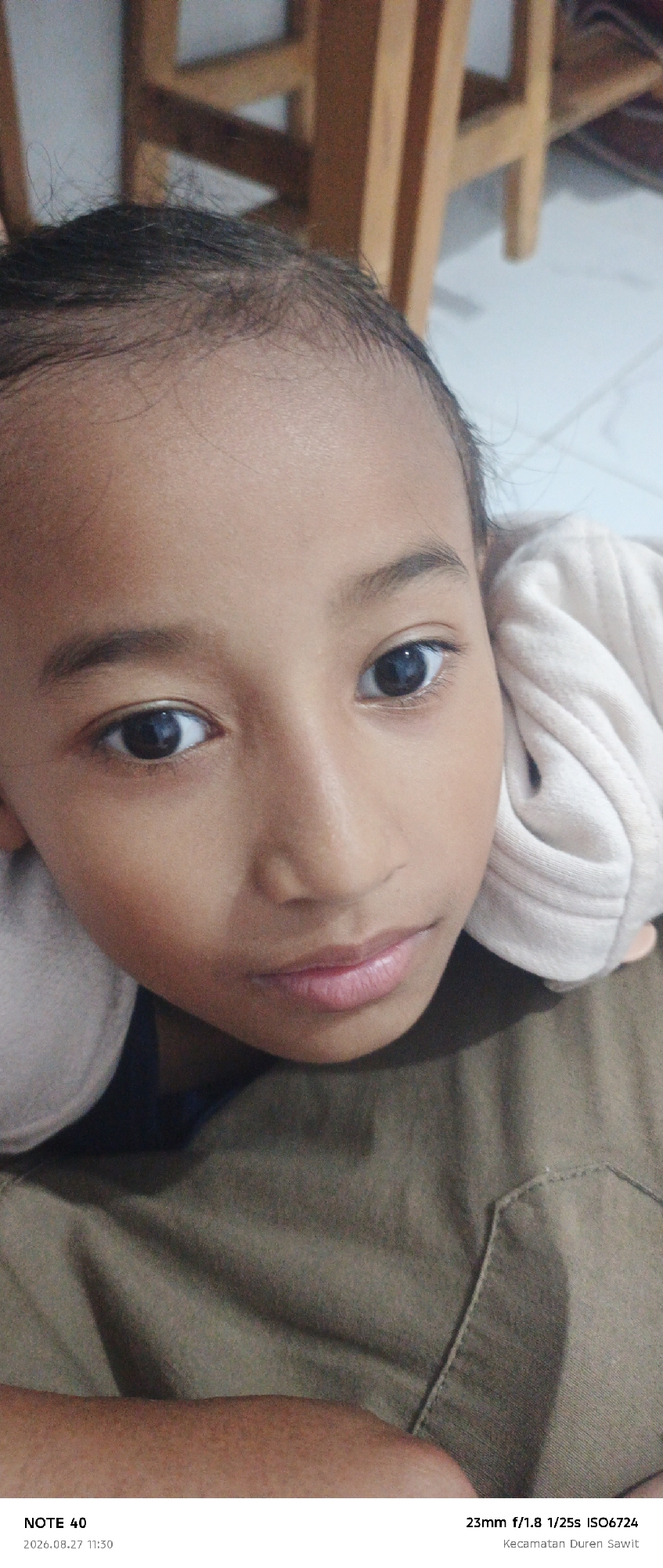 The image is a close-up, eye-level, portrait-style photograph of a young girl's face. She is looking directly at the camera with a neutral expression. Her eyes are dark, and her eyebrows are slightly arched. Her lips are closed and appear to have a subtle pink hue. Her skin is a warm, medium tone. She has dark, fine hair that is slightly messy around her forehead and temples.
She is wearing a light-colored garment, possibly a hoodie or sweatshirt, around her shoulders and head. The background is blurred, but it appears to be an indoor setting with some wooden furniture and a tiled floor. The lighting is soft, illuminating her face.
At the bottom of the image, there is text providing photographic information: "NOTE 40 2026.08.27 11:30 23mm f/1.8 1/25s ISO6724 Kecamatan Duren Sawit". This indicates the photo was taken on August 27, 2026, at 11:30 AM, with a 23mm lens at an aperture of f/1.8, a shutter speed of 1/25s, and an ISO of 6724. "Kecamatan Duren Sawit" is a subdistrict within the city of East Jakarta, Indonesia, suggesting the location where the photograph was taken.