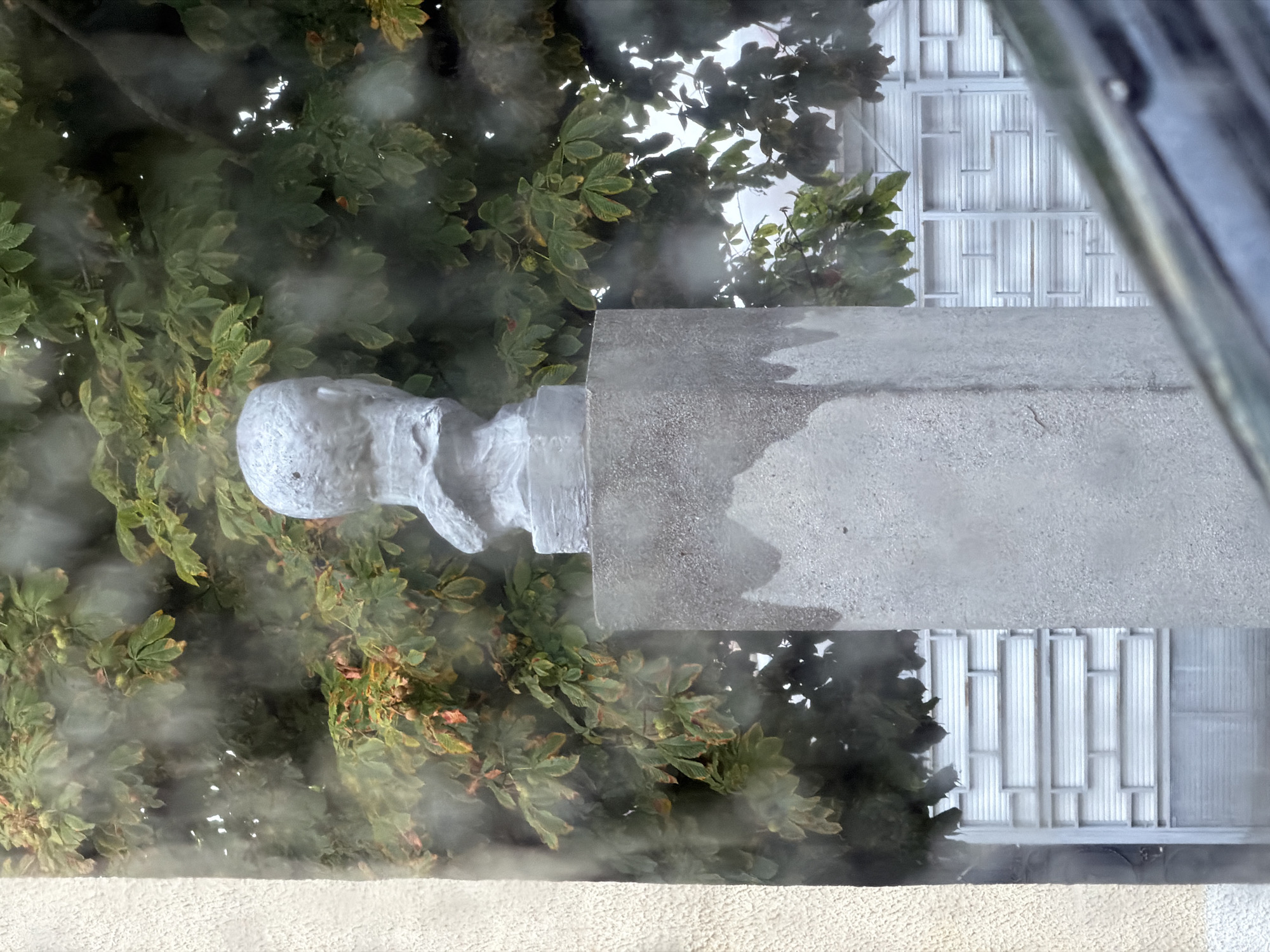 The image depicts a bust of a person placed on top of a concrete pedestal. The bust is made of a light-colored material and appears to be weathered. The pedestal is tall and rectangular, with a rough texture. Behind the bust and pedestal, there is a dense canopy of green leaves from a tree, some of which show signs of autumn discoloration. Through the leaves and partially obscured by what appears to be steam or mist, a building with horizontal white panels is visible. The overall scene suggests an outdoor setting, possibly in a park or a courtyard, with the mist adding a slightly obscured and atmospheric quality. The location is specified as Cherven, Belarus, but no specific landmarks are identifiable from the image itself.