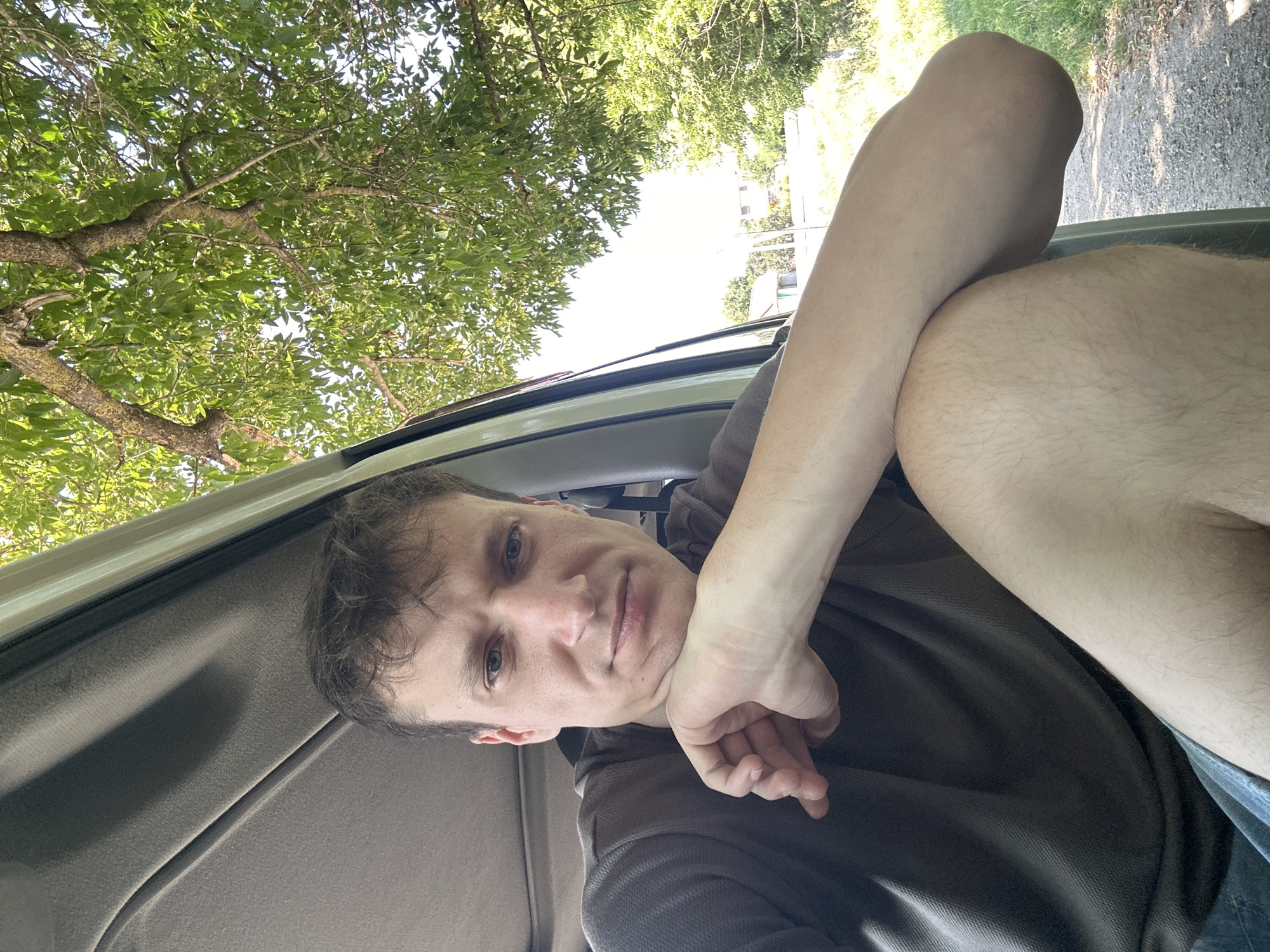 A man is seated in the interior of a vehicle, his right arm resting on his bent leg. He looks directly at the camera with a neutral expression, his head supported by his hand. The vehicle's interior is visible, with a grey fabric headliner and a portion of the door frame. Outside the vehicle, through the open door and window, lush green trees and foliage are prominent, suggesting a natural, possibly rural, setting. The ground appears to be gravel. The image is a selfie, taken from a slightly low angle. There is no indication of the specific location within Makiv, Ukraine.