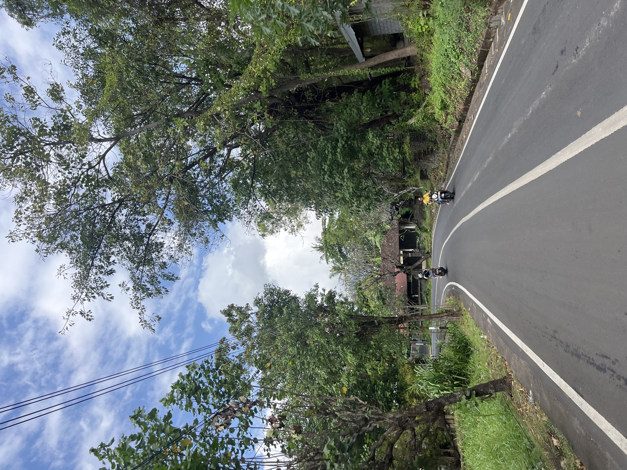 A road in Abang, Indonesia, is flanked by lush green trees and vegetation. The sky is a mix of blue and white clouds. Two individuals are riding scooters on the road, one in front of the other. The road is paved and has a white line dividing the lanes. There are houses visible in the background, with traditional Indonesian architecture. The overall scene depicts a peaceful and scenic rural landscape.