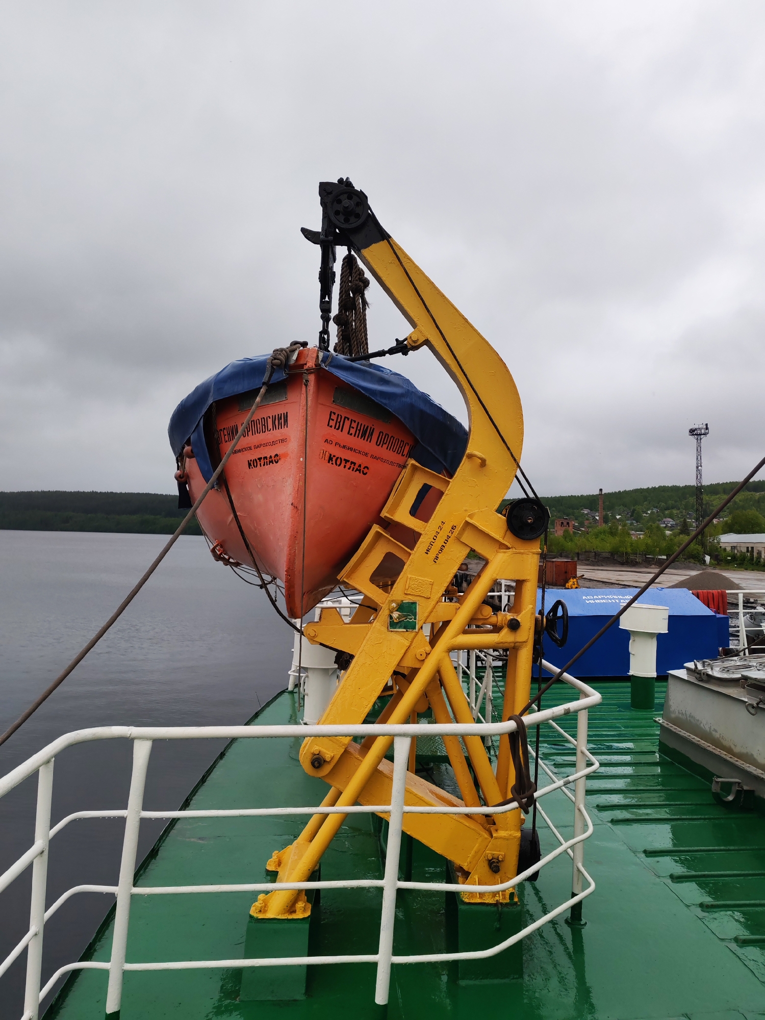 The image captures a bright orange lifeboat suspended from a yellow davit on the port side of a ship, against an overcast sky. The ship's green deck appears wet and reflective, suggesting recent rain or high humidity. This scene is likely set on a large body of water, possibly Lake Onega near Medvezhyegorsk, Russia, given the background of dense green forest covering hills.
The lifeboat is partially covered with a blue tarpaulin and features prominent Cyrillic text. On its bow, the name "ЕВГЕНИЙ ОРЛОВСКИЙ" (Yevgeny Orlovsky) is clearly visible, along with "АО РЫБИНСКОЕ ПАРОХОДСТВО" (JSC Rybinsk Shipping Company) and "КОТЛАС" (Kotlas), which likely identify the lifeboat's name, owner, and homeport, respectively. The yellow davit itself has codes "ИСП.04.24." (presumably "Inspection 04.24.") and "ПРОВ.04.26." (likely "Verification 04.26."), possibly indicating maintenance or testing dates. Further down the ship's deck, a blue cover bears the text "АВАРИЙНЫЙ ИНВЕНТАР" (Emergency inventory).
The davit structure is robust, equipped with black pulleys, chains, and thick ropes that secure the lifeboat. A white metal railing runs along the edge of the ship's deck. In the distance, beyond the calm water, a forested shoreline stretches, with a glimpse of an industrial or port area on the right. This includes a tall metal communication tower, what appears to be an old brick factory chimney, and other buildings. The overcast sky indicates a cloudy daytime, providing soft, diffused lighting to the tranquil scene.